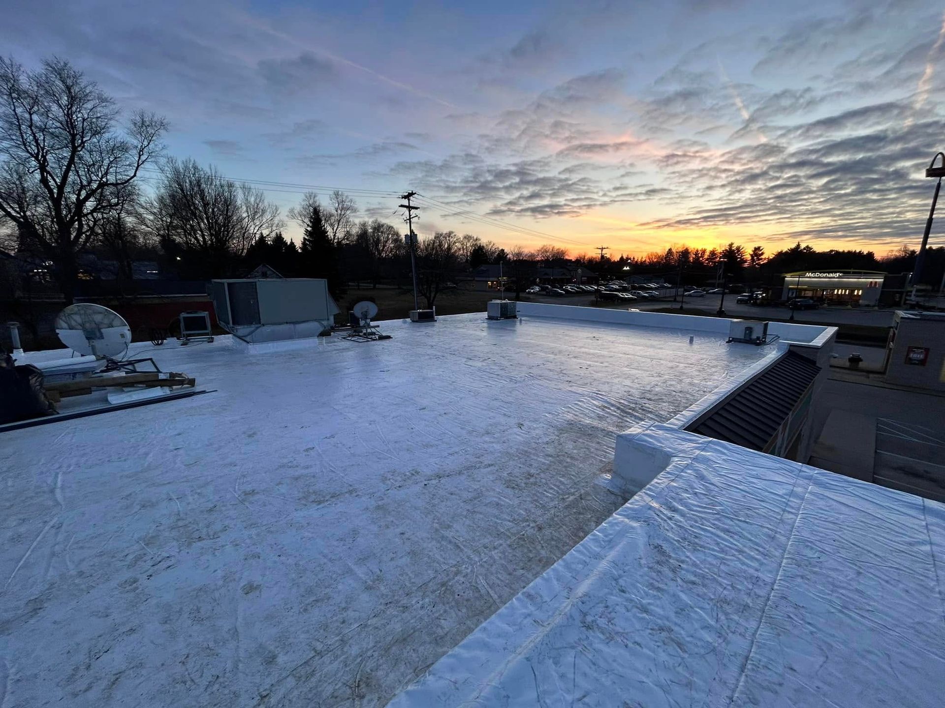 Snow-covered rooftop at dawn with a colorful sunrise and bare trees in the distance