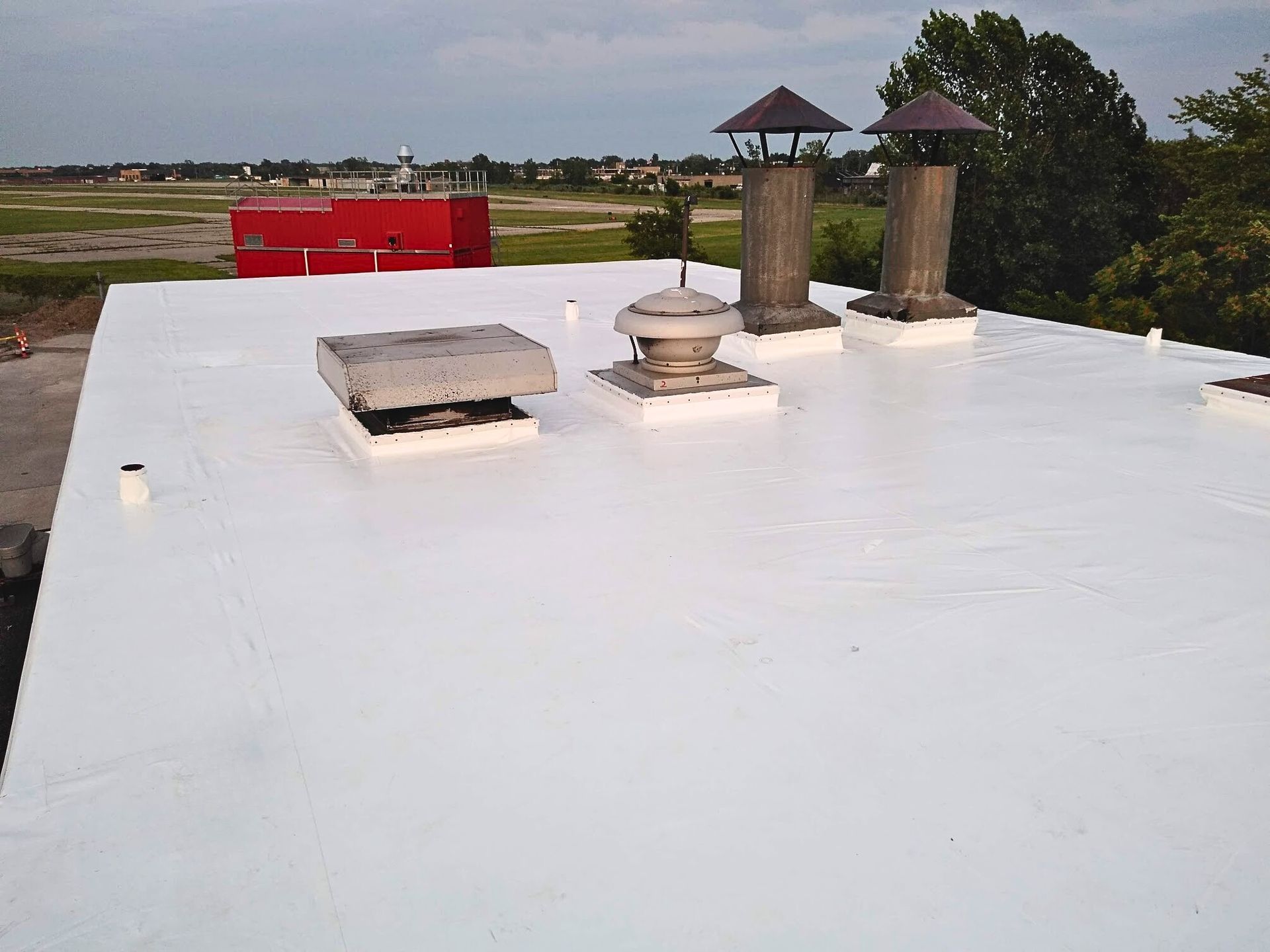 Rooftop with white waterproof coating, vents, and two chimney stacks against a rural landscape