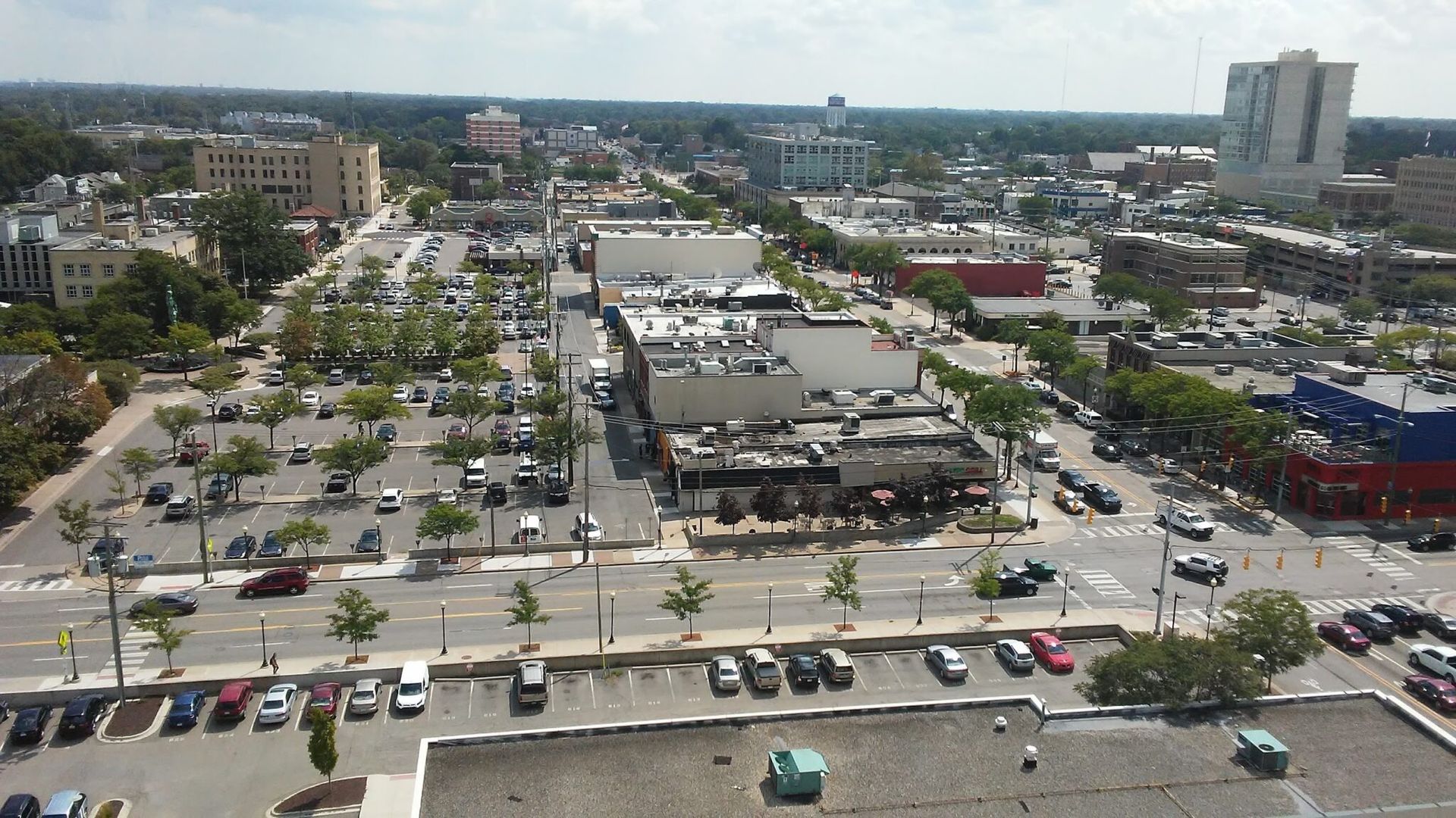 Aerial view of a busy urban intersection with parking lots, storefronts, and mid-rise buildings under a clear sky