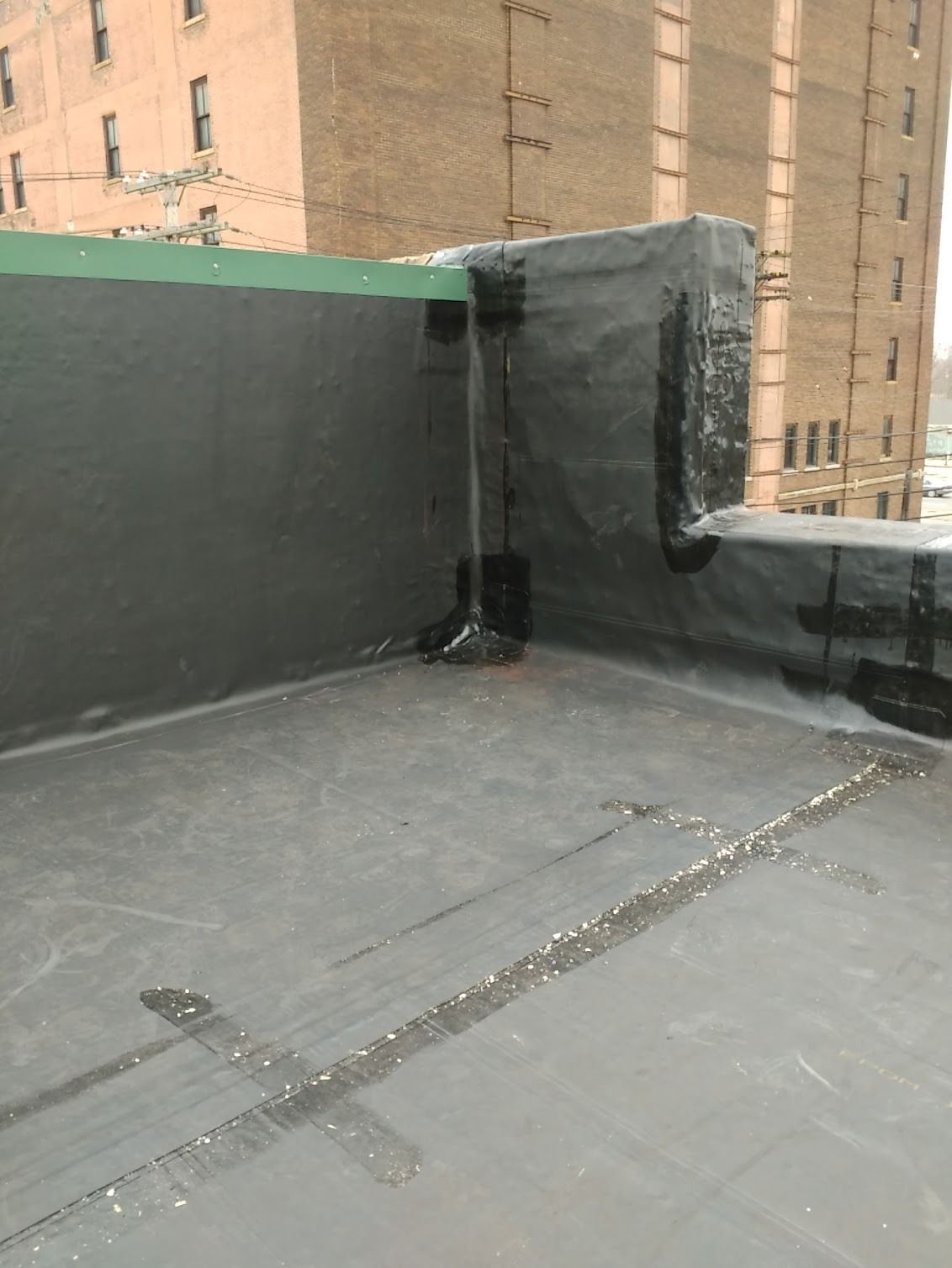 Rooftop corner with gray waterproofing, green parapet wall, and tan brick buildings in the background
