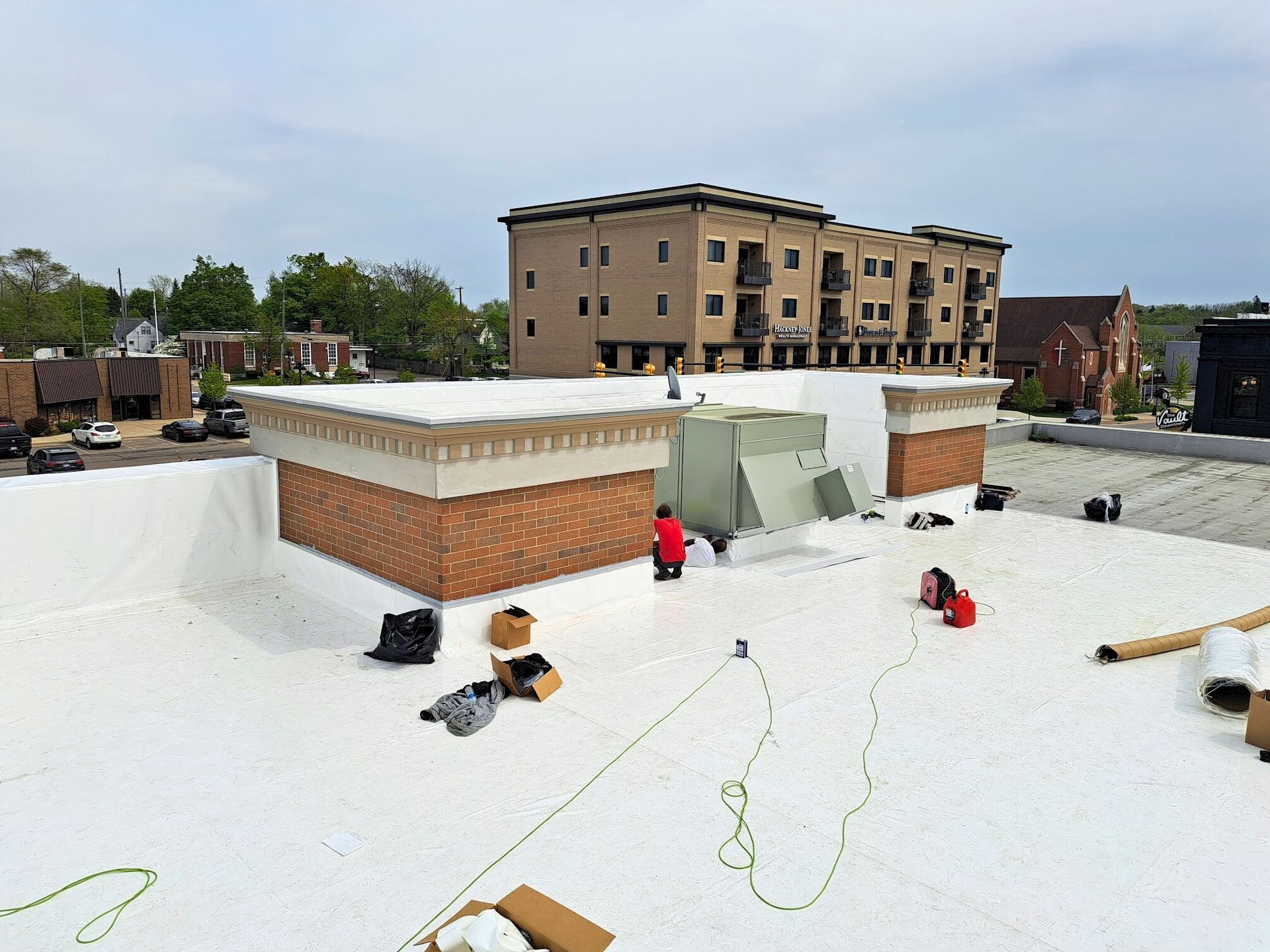 Flat white roof with workers and tools beside a brick rooftop structure under a cloudy sky