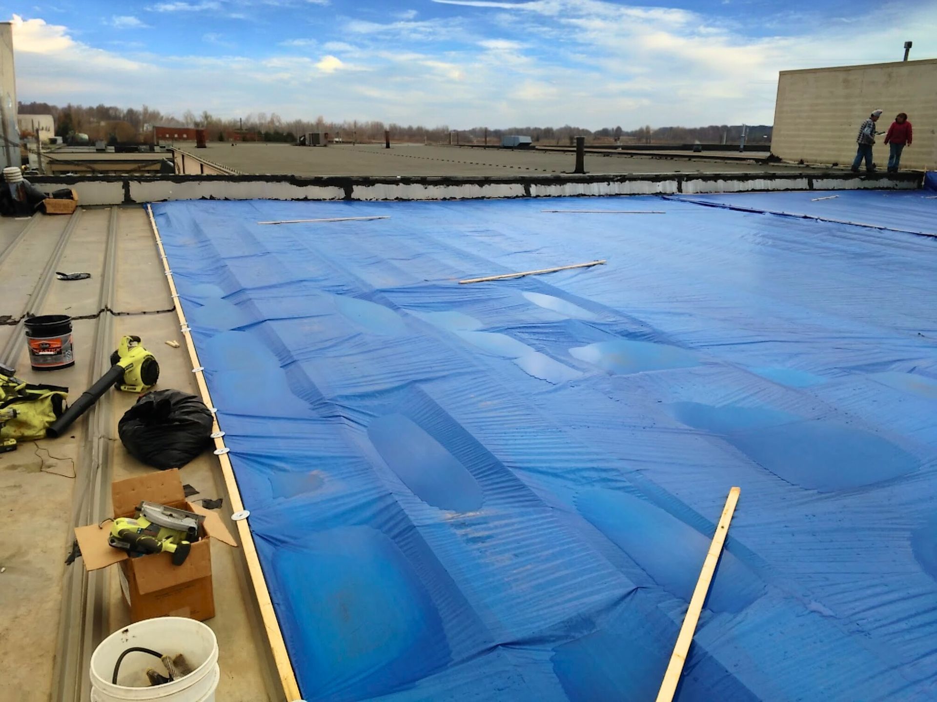 Workers covering a rooftop pool with a large blue tarp under a cloudy sky