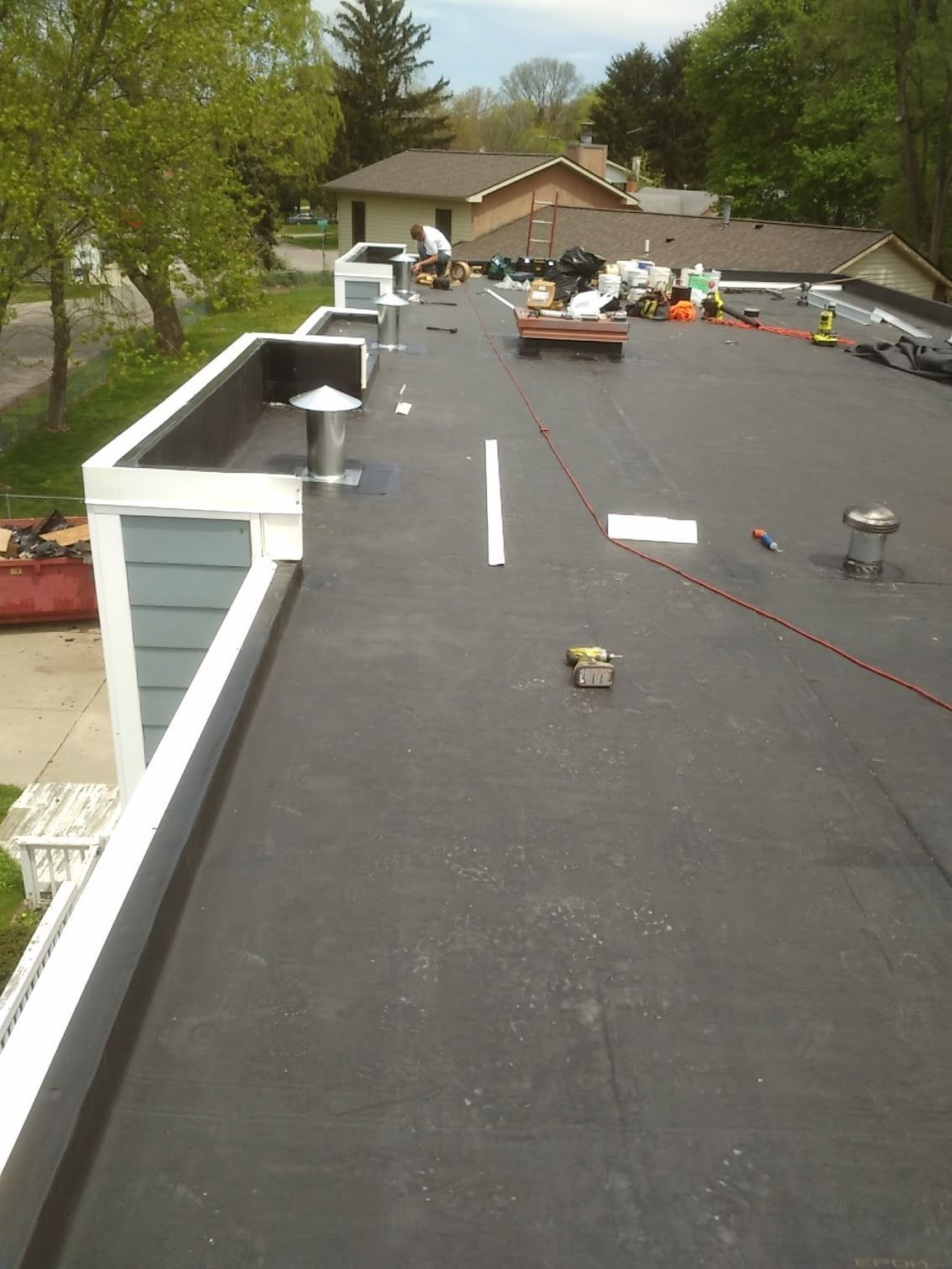 Flat black rooftop under construction with tools and materials, trees and a house in the background