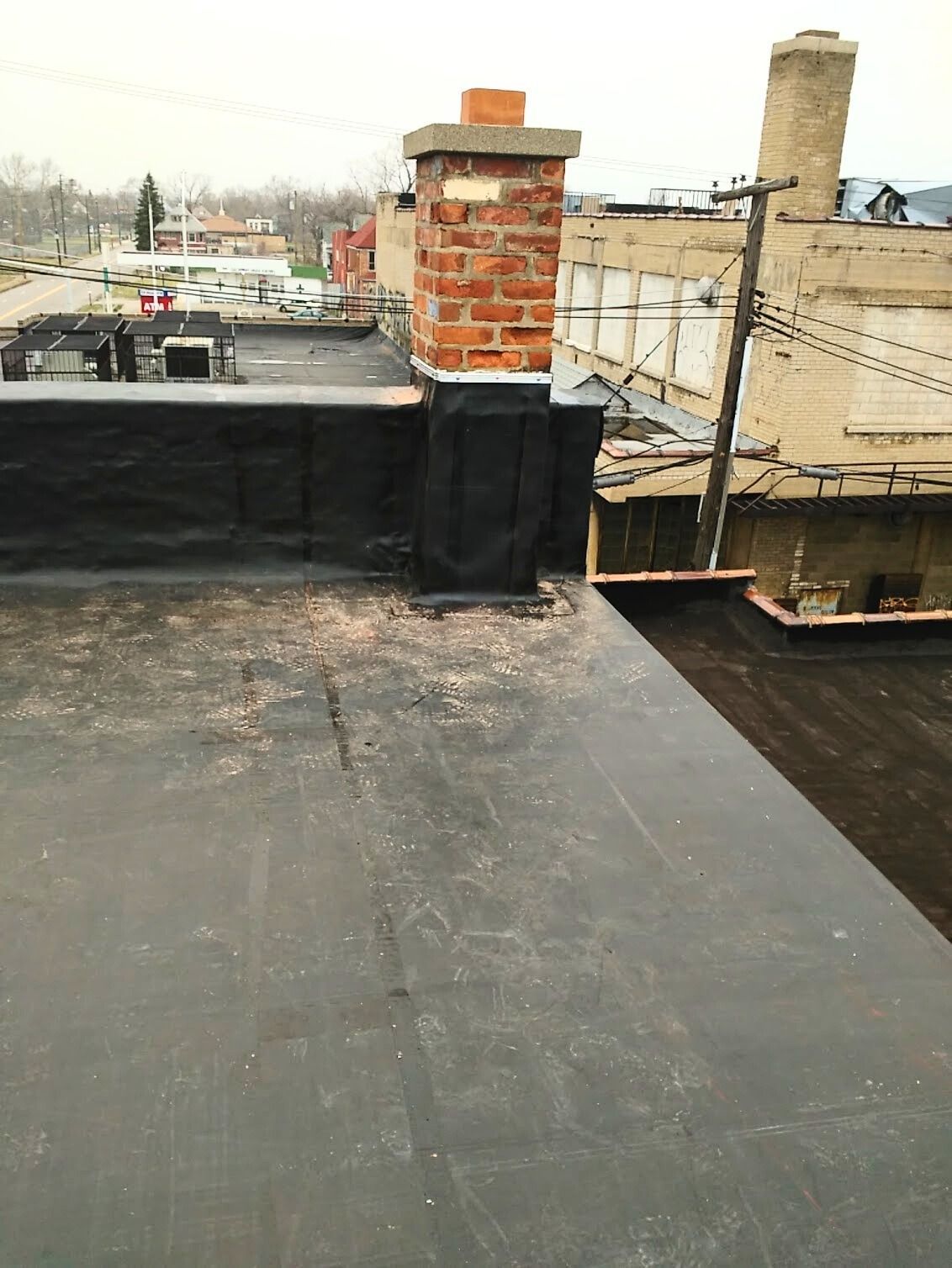 Flat rooftop with black waterproofing, a brick chimney, and neighboring buildings under a cloudy sky