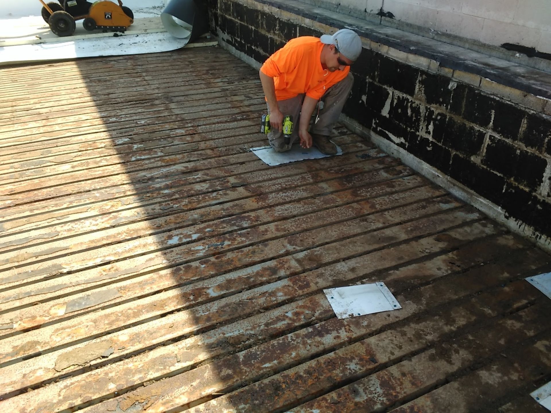 Worker in orange jacket kneels on a rusty metal deck, inspecting the surface near a wall.