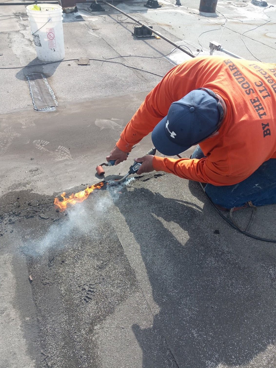 Worker in orange safety vest using a torch to repair a roof seam with flames visible.