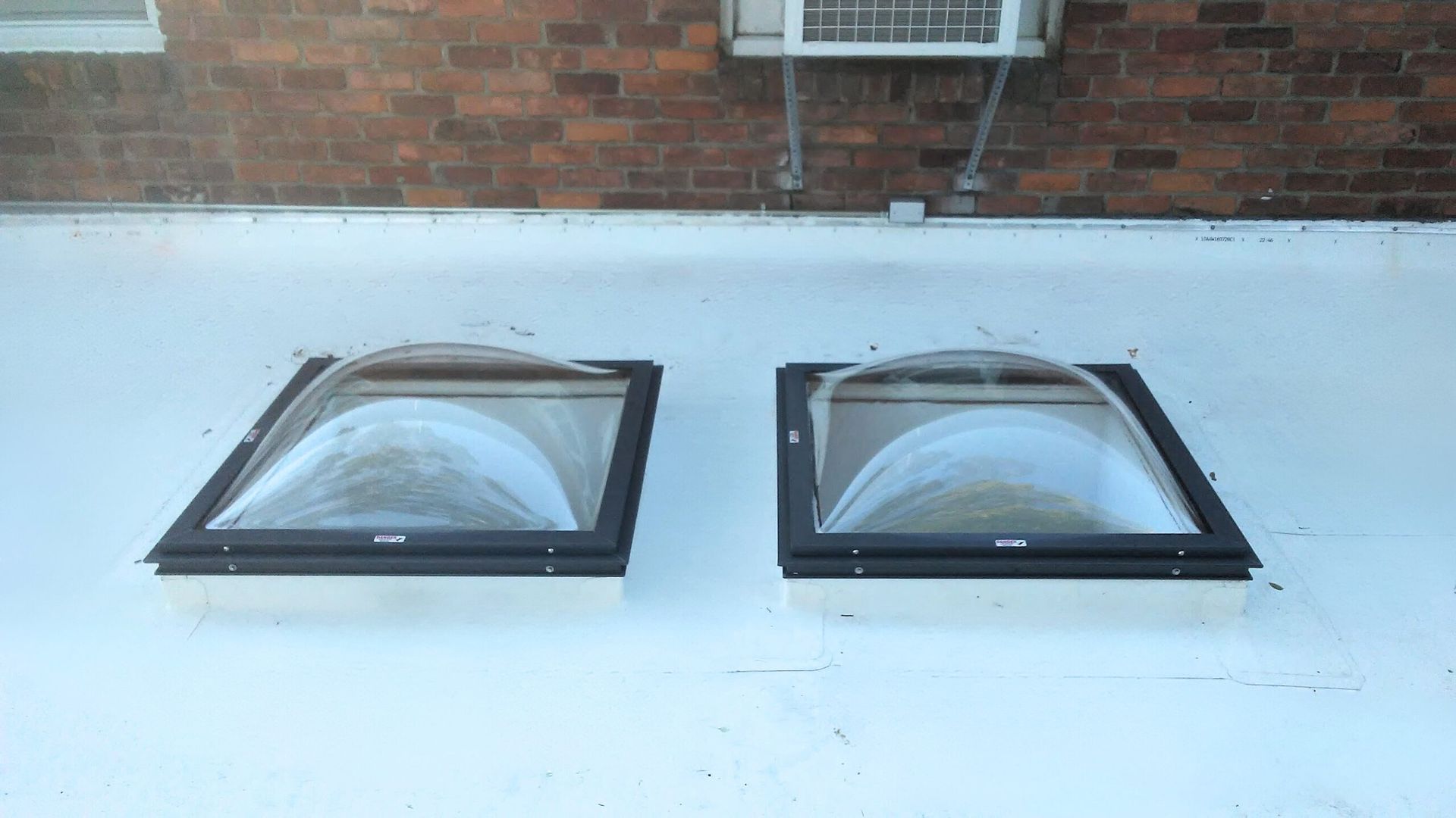 Two black-framed skylights on a white flat roof beside a brick wall.