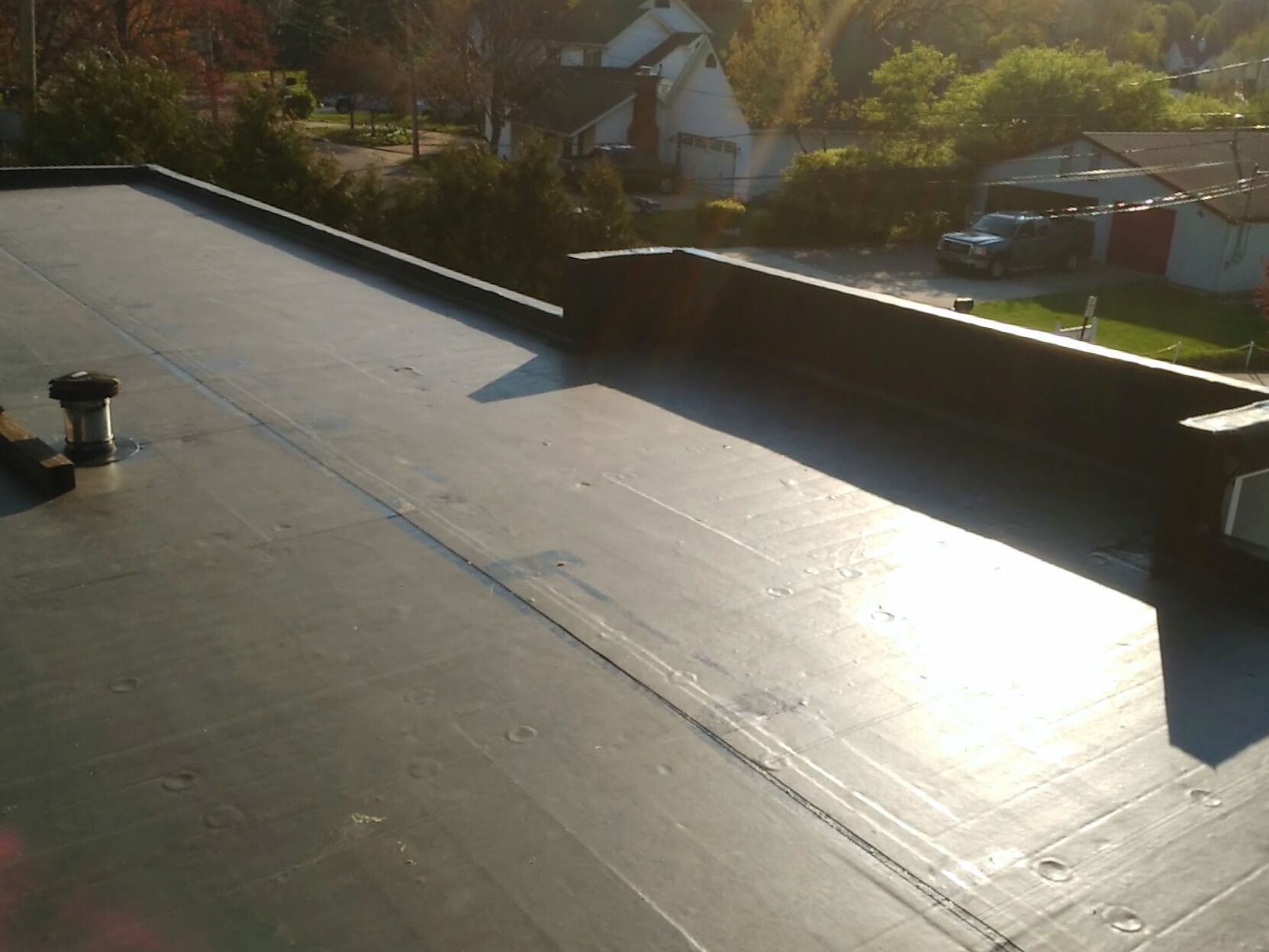 Flat black rooftop with a vent, sun glare, and houses in the background