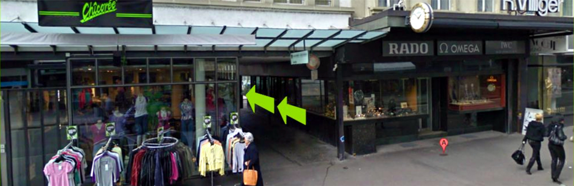 Geschäfte mit Kleidung an Kleiderständern und Rado Omma Uhren, Straßenansicht. Grüne Pfeile weisen auf den Durchgang hin.