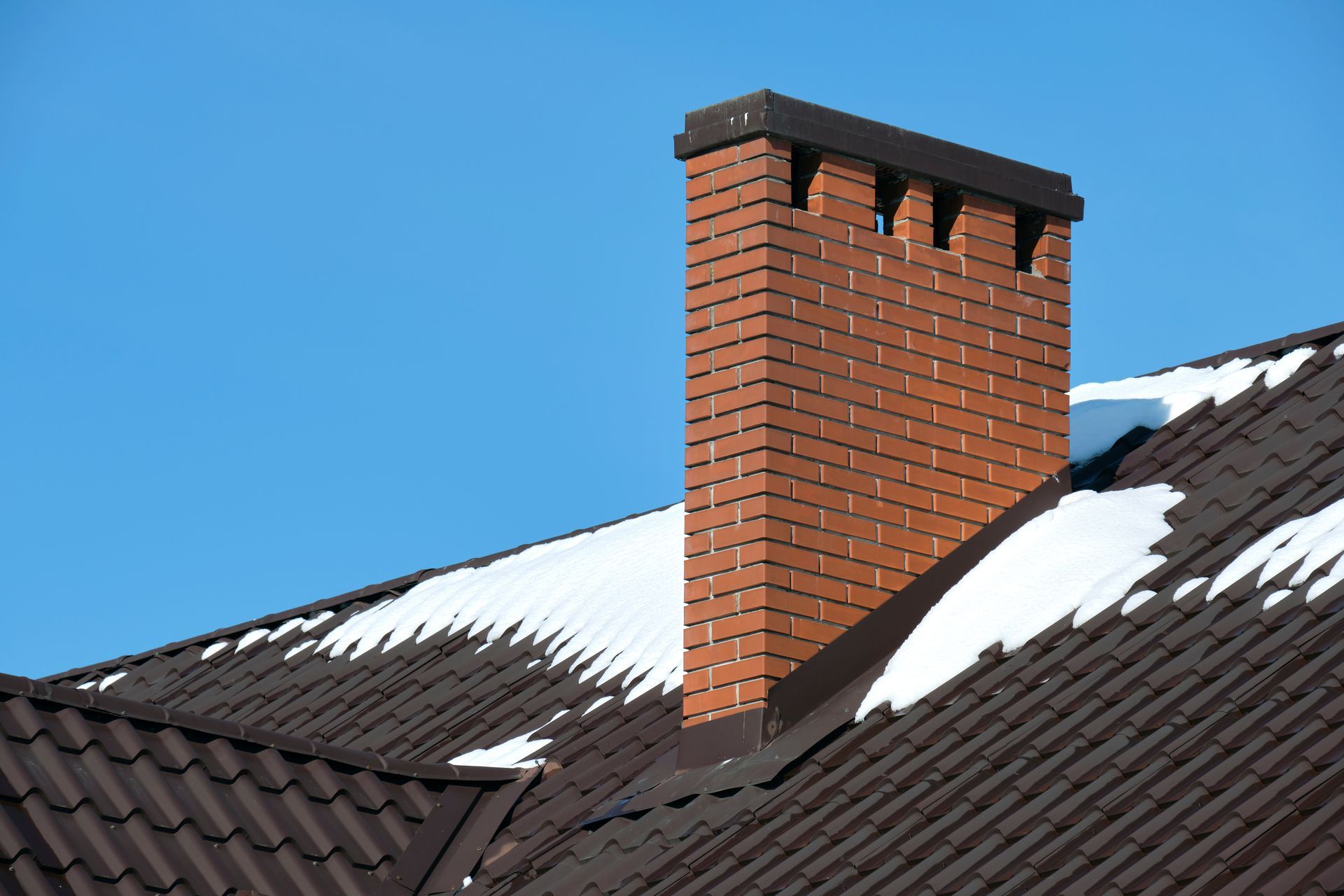 Brick chimney on a snow-covered dark roof under a blue sky.