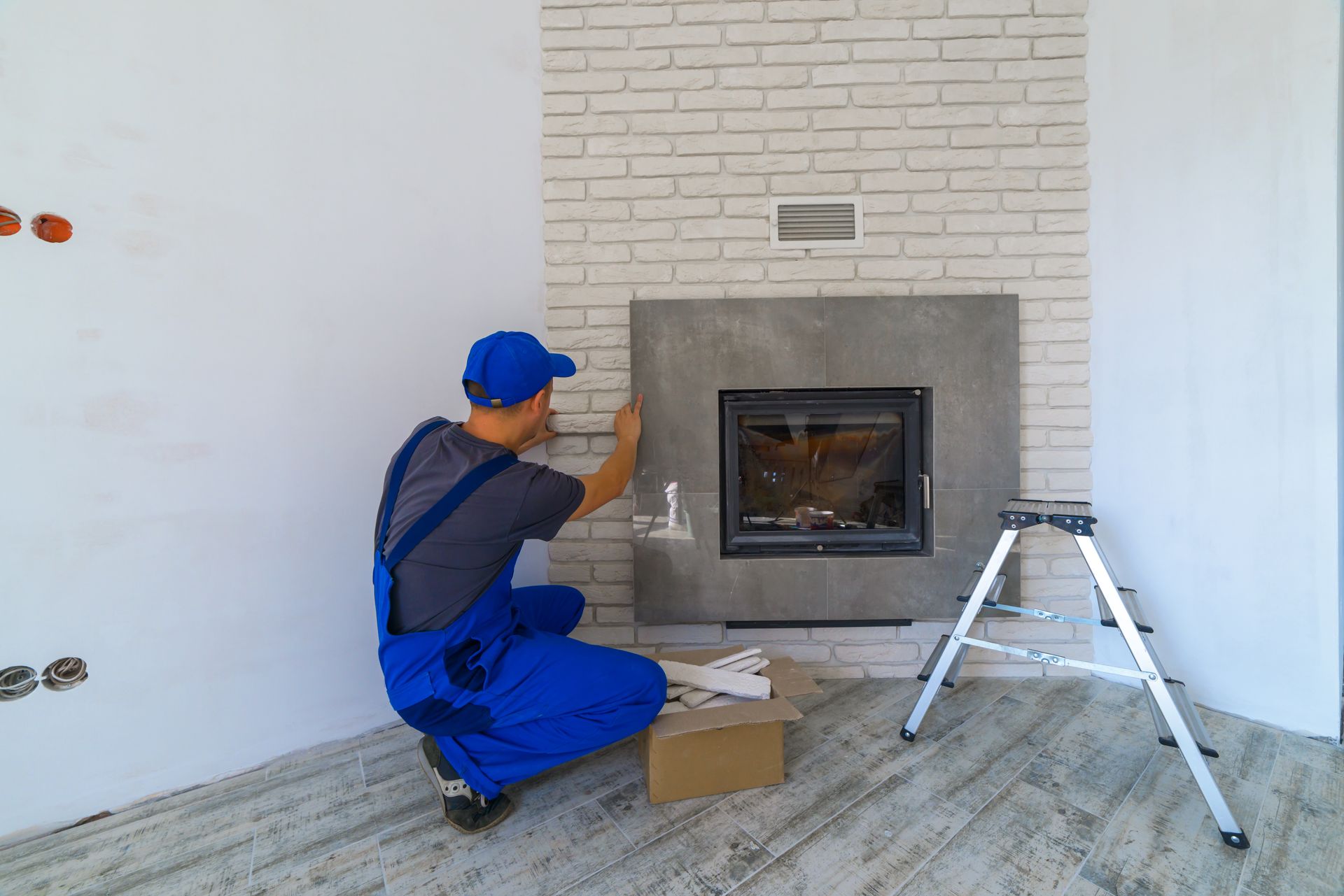 Worker in blue overalls installing a fireplace in a bright living room, with a step ladder nearby.