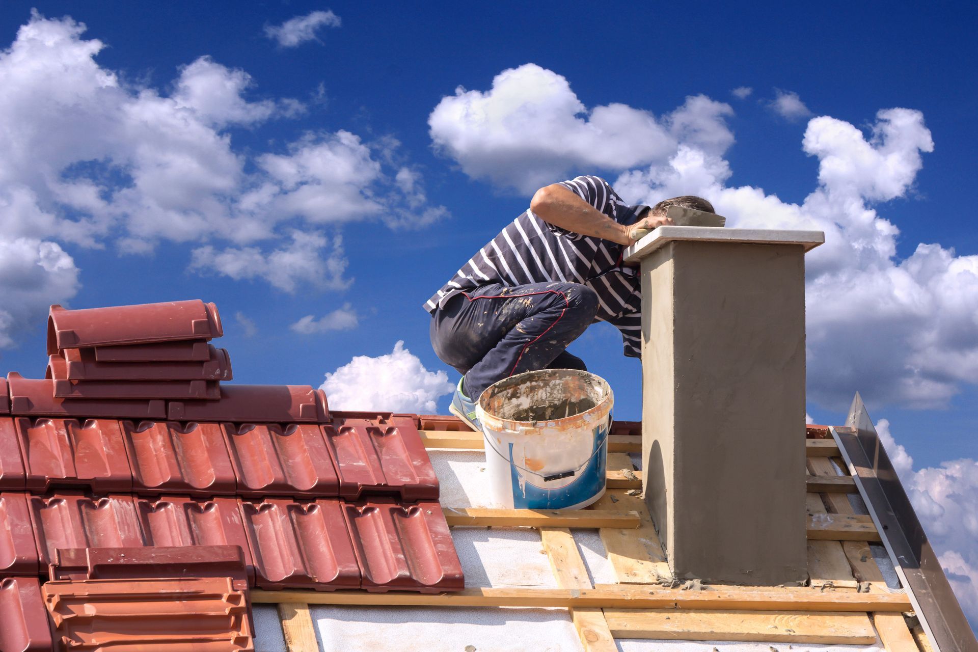 Worker on a tiled roof, kneeling beside a chimney under a blue sky with clouds.