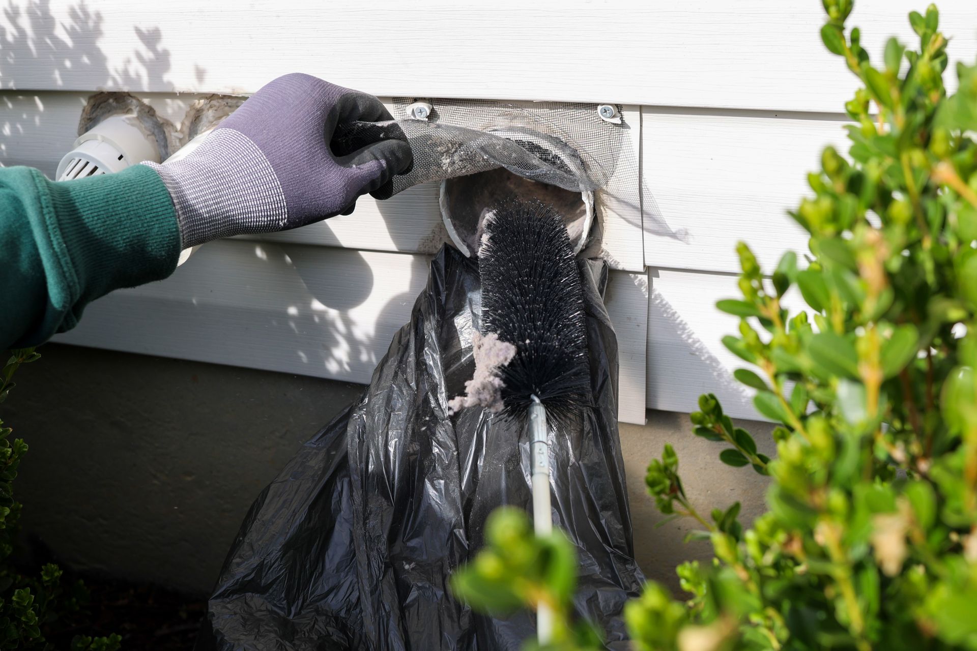 Gloved hand scraping peeling paint from white house siding under an exterior window.