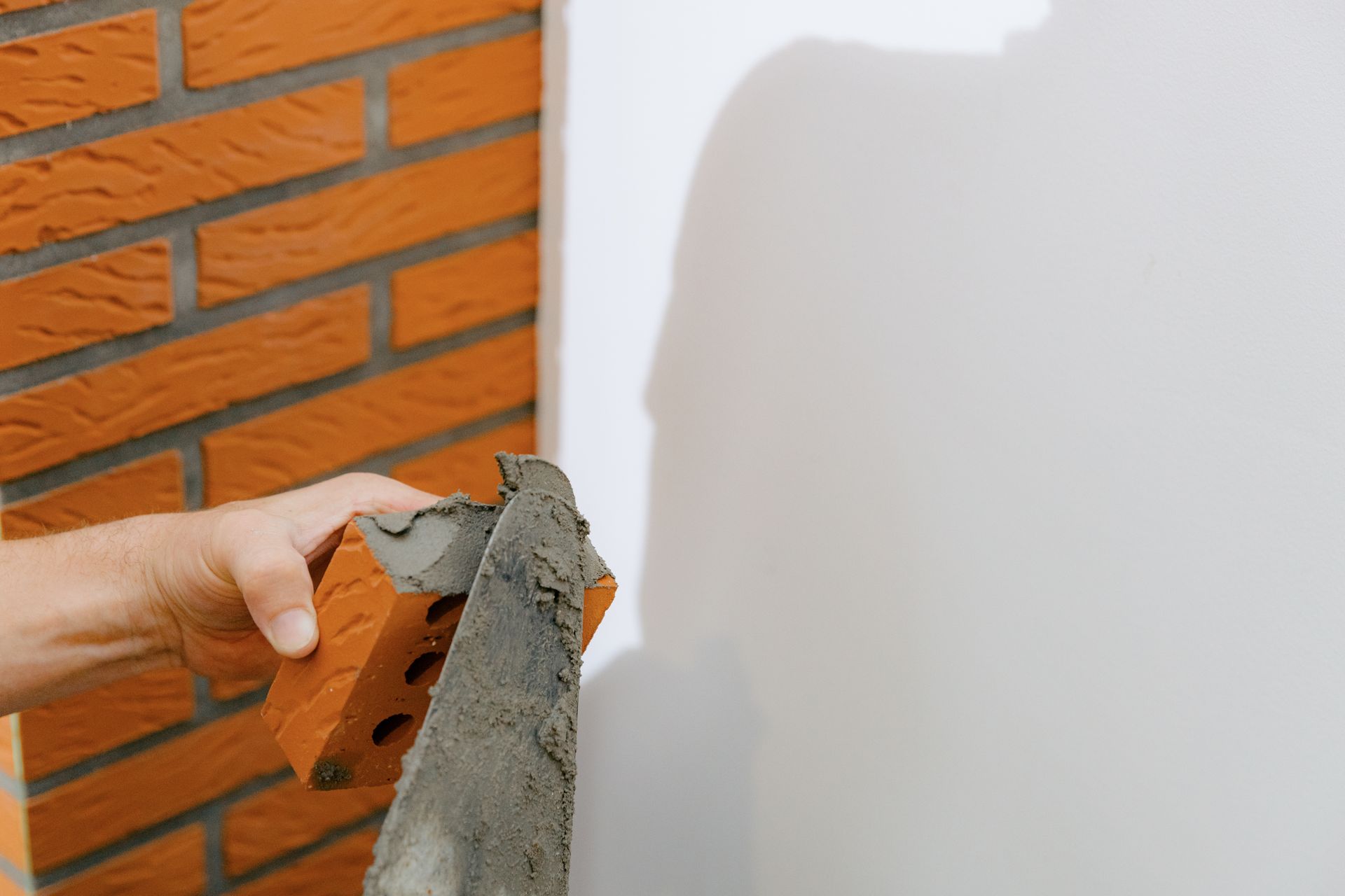 Hand trowel spreading wet plaster on a white wall beside orange brick siding. Hand trowel spreading wet plaster on a white wall beside orange brick siding.