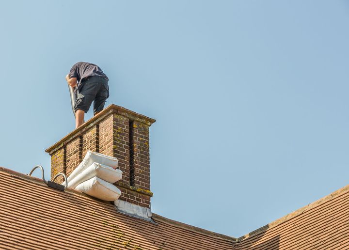 Worker standing on a brick chimney atop a tiled roof under a clear blue sky.