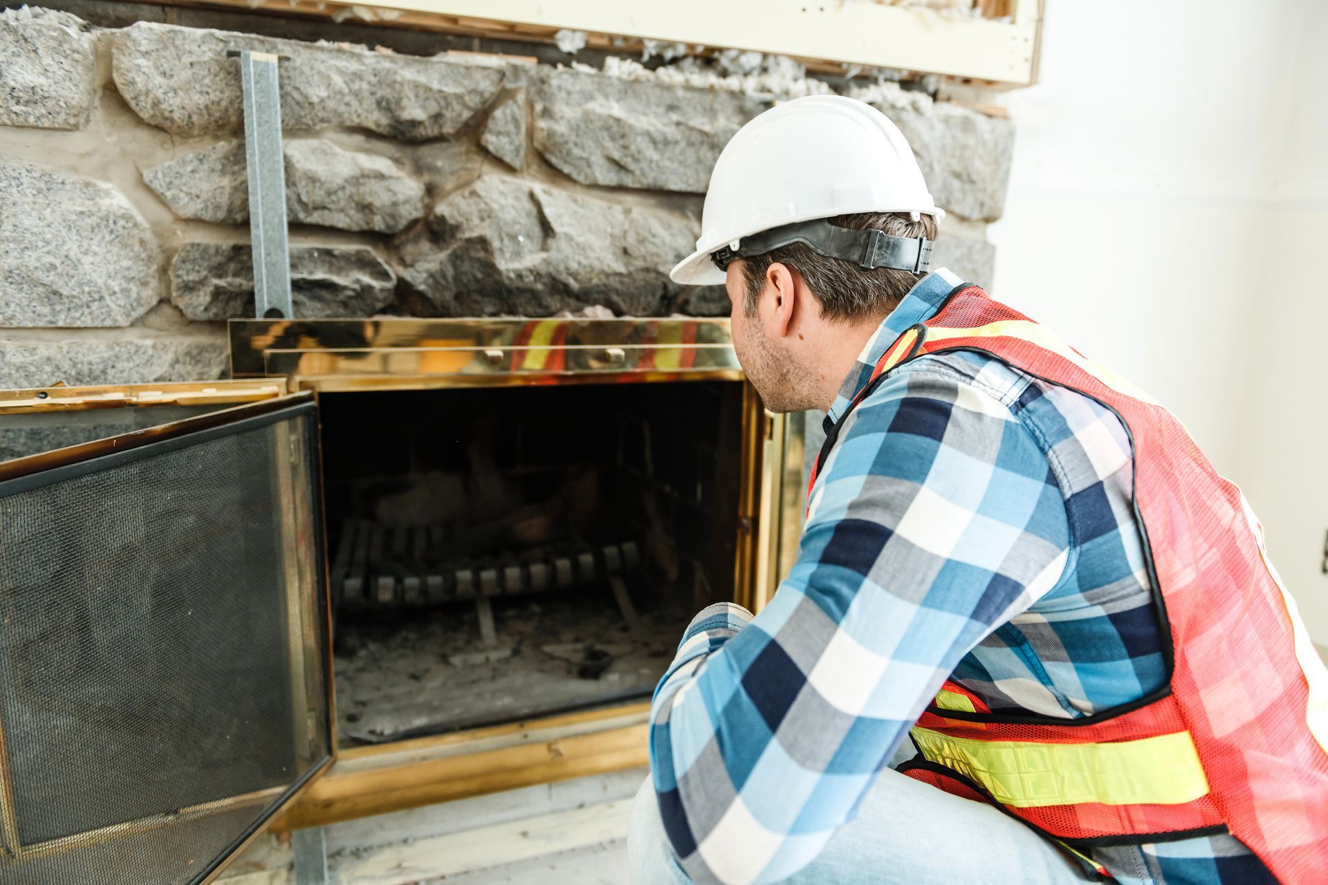 Worker in hard hat inspecting a fireplace or furnace opening inside a stone-walled room.