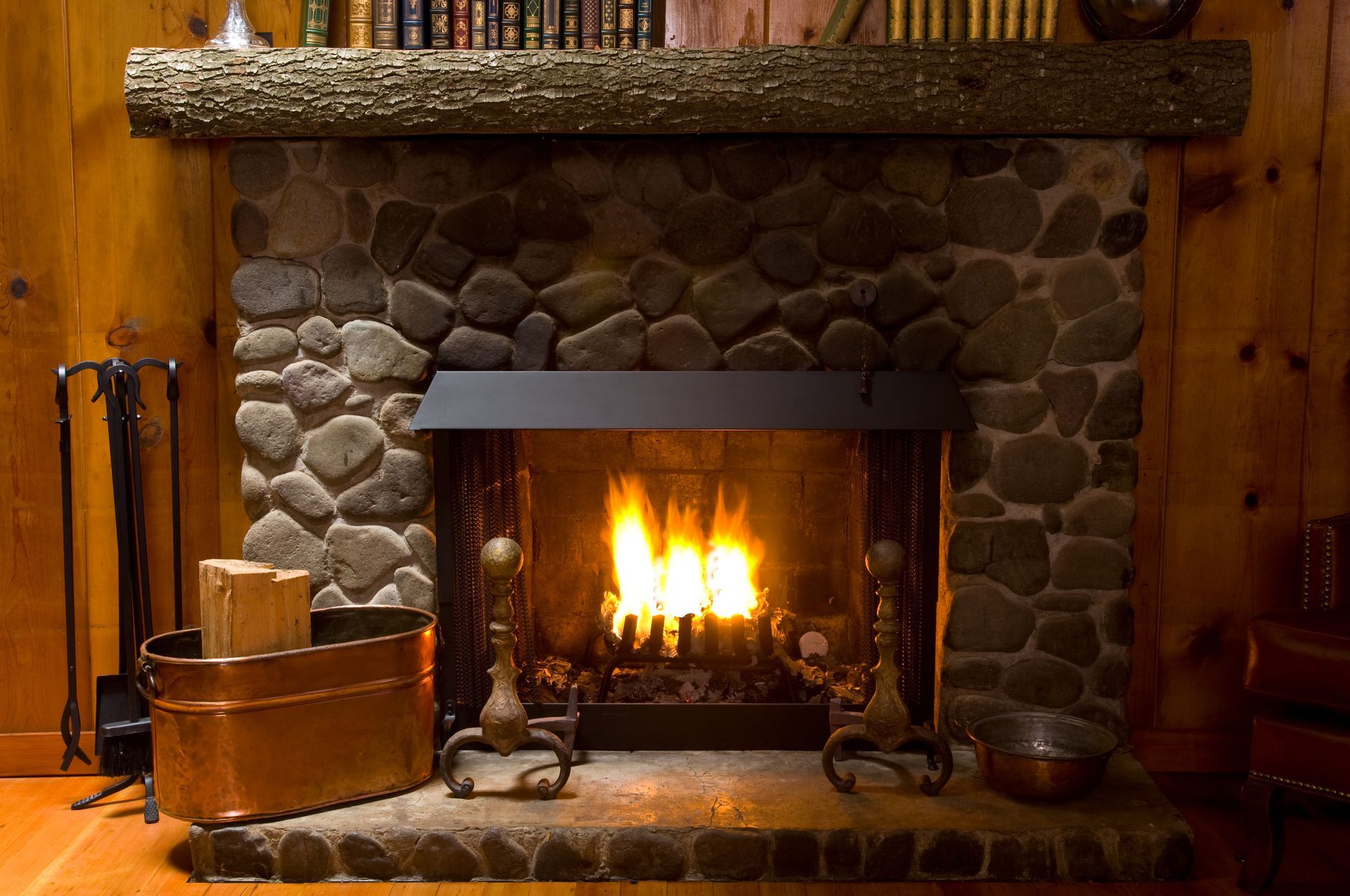 Stone fireplace with a burning fire, dark mantel, and wooden hearth accessories in a cozy room. Stone fireplace with a burning fire, dark mantel, and wooden hearth accessories in a cozy room.