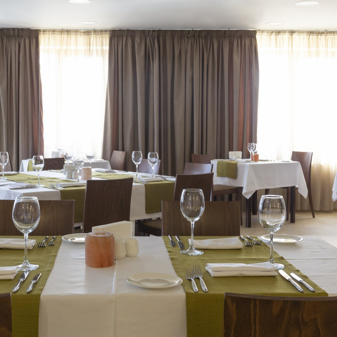 Commercial dining room with set tables with empty wine glasses, backed by half open natural drapes letting sunlight filter through sheers.