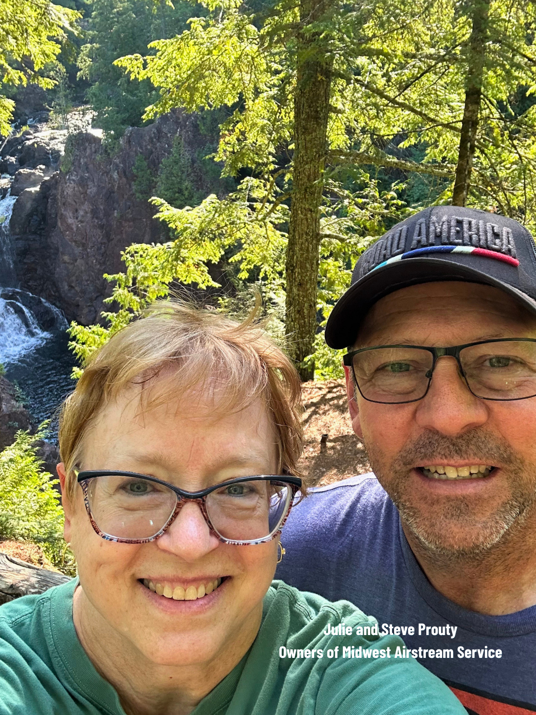 Smiling couple taking a selfie in front of a waterfall and sunlit trees.