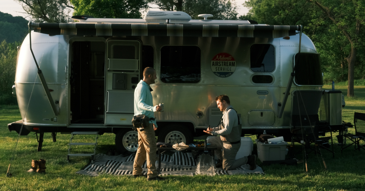 Two men, outside an Airstream travel trailer, cooking.
