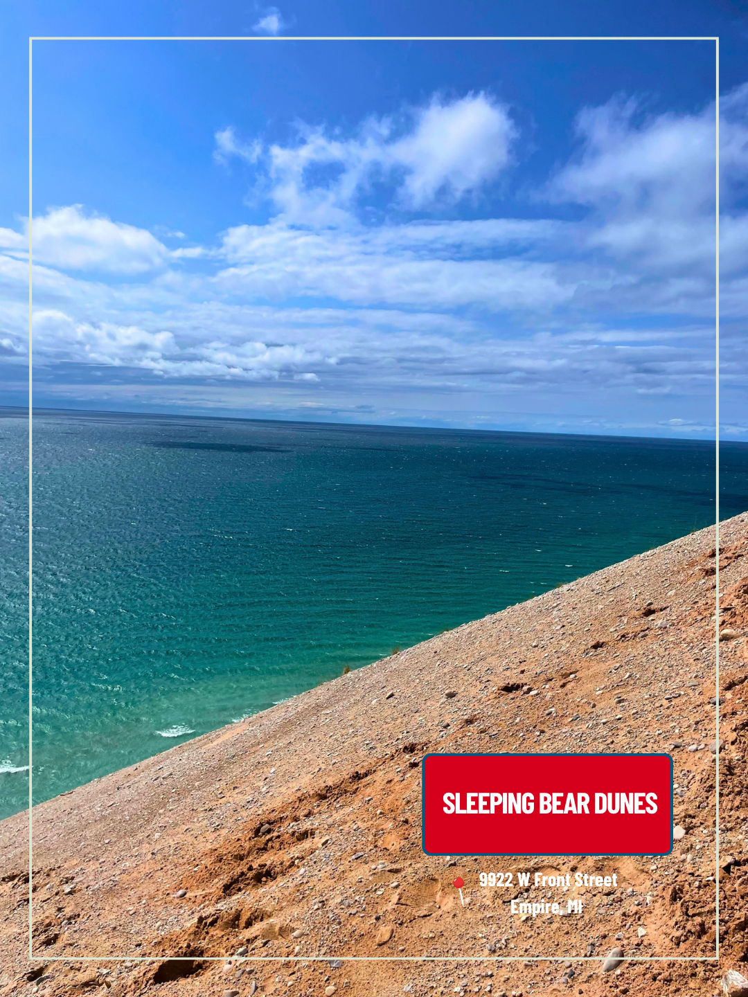 Turquoise ocean beside a sandy dune under a blue cloudy sky, with a red “Sleeping Bear Dunes” sign
