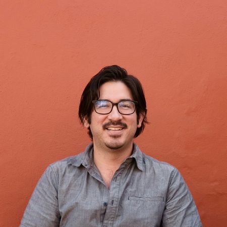 A man in a grey collared shirt with black glasses and a gentle smile