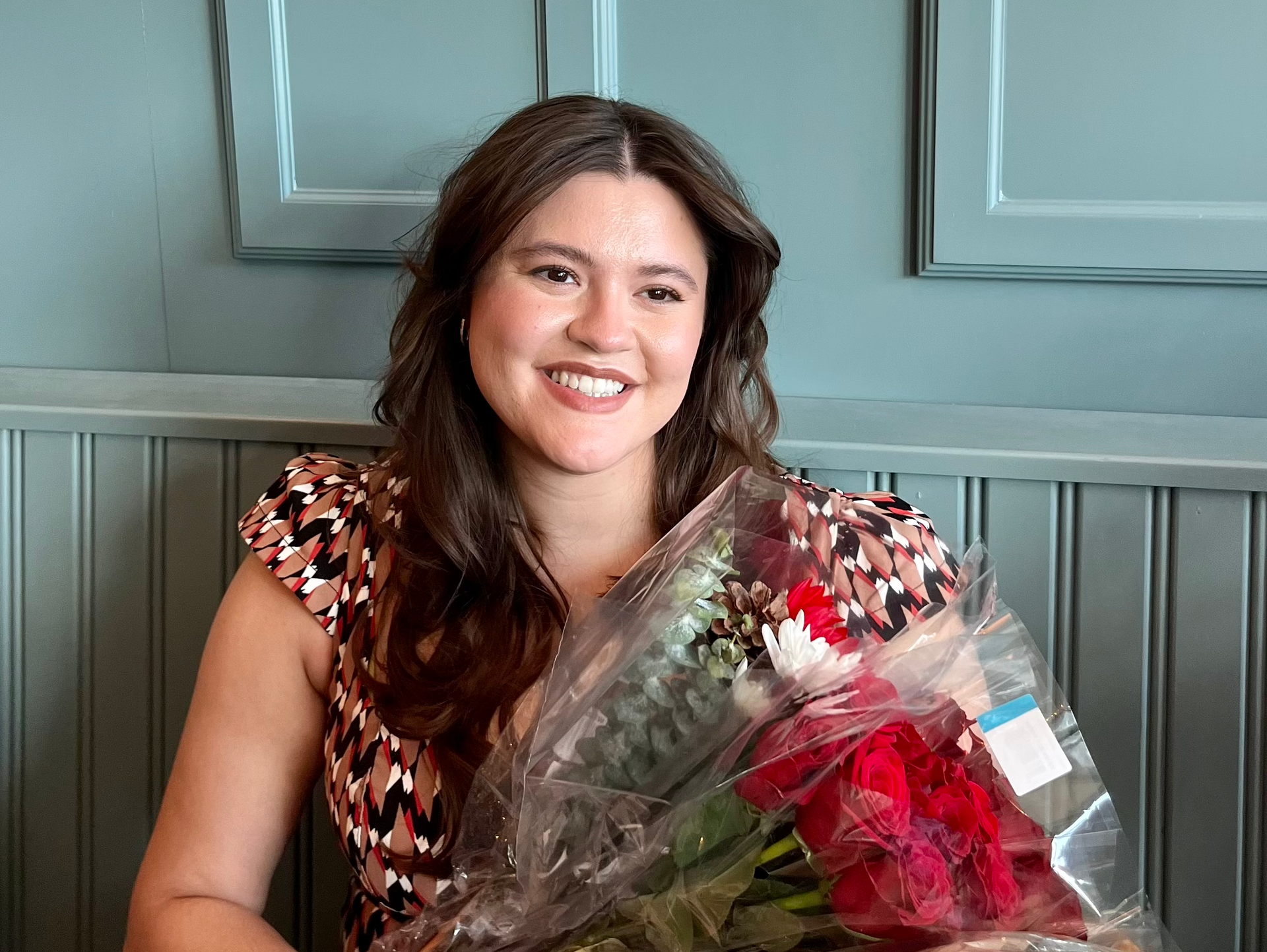 A woman in a red tank top is smiling in front of a bush of flowers.