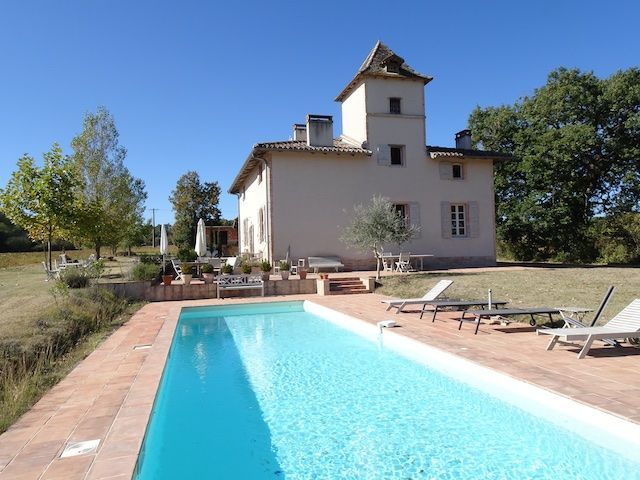 A long, rectangular swimming pool in front of a cream-colored house with a small tower. Sunny day.