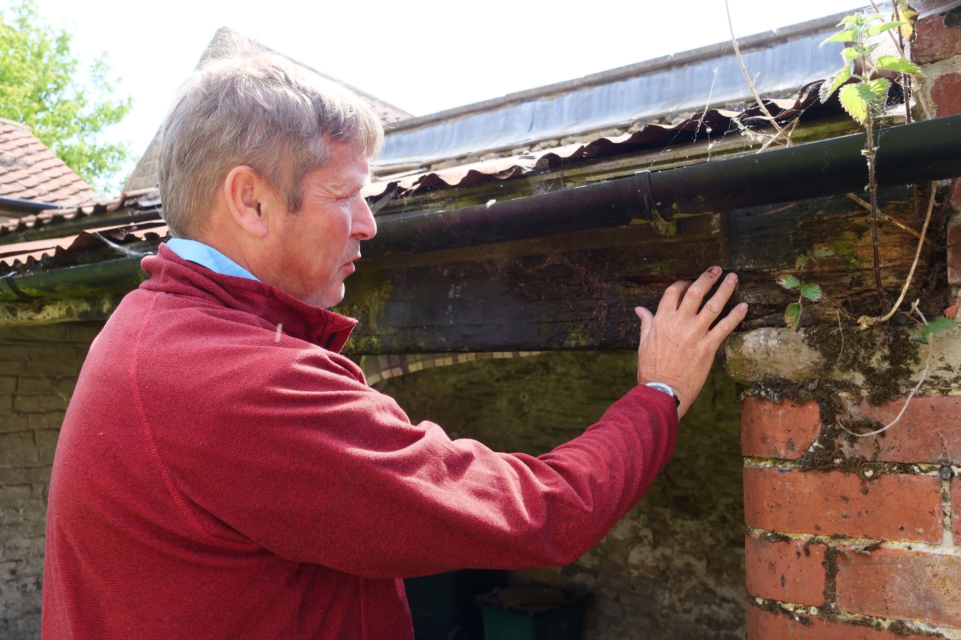 Man in red jacket examining a deteriorating brick building and gutter, outdoors on a sunny day.