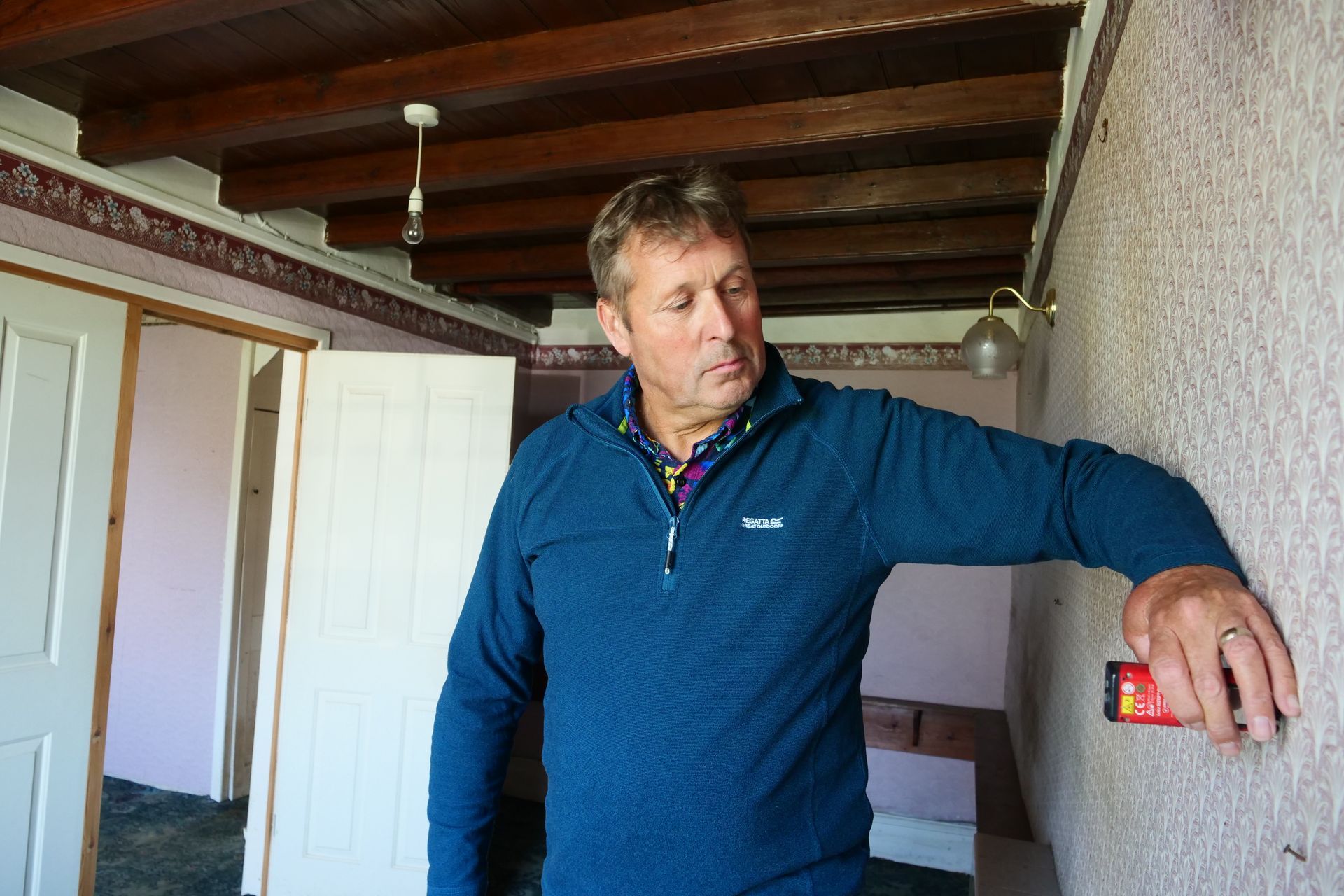 Man in blue sweater using a stud finder on a textured beige wall in a room with exposed wood beams.