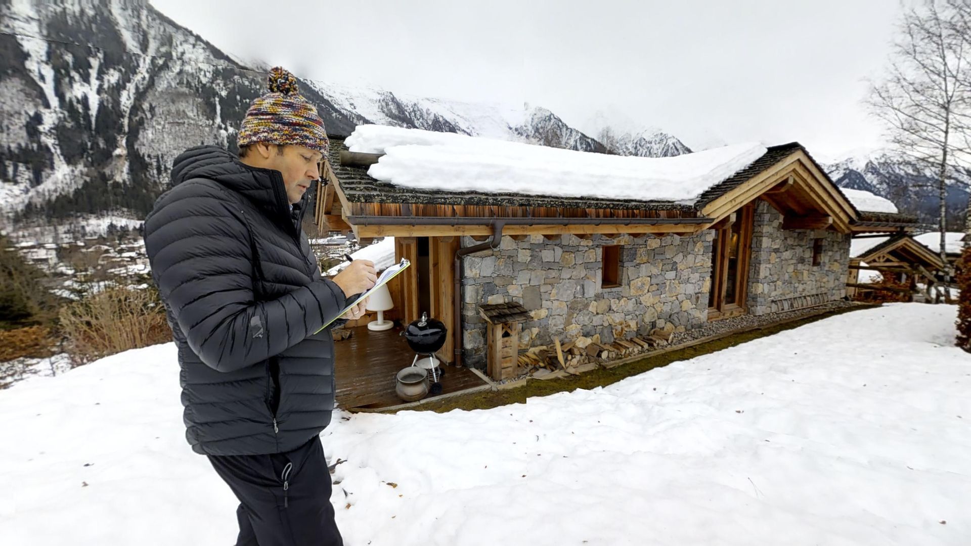 Woman in winter clothes using a phone outside a stone cabin covered in snow, with snow-covered mountains in the background.