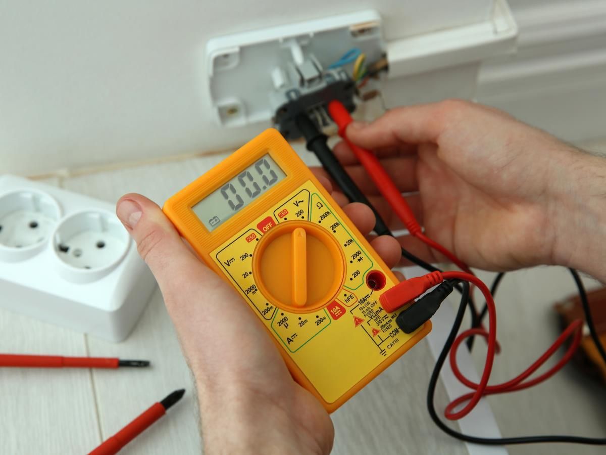 Hands testing an exposed wall outlet with a yellow multimeter and red probes