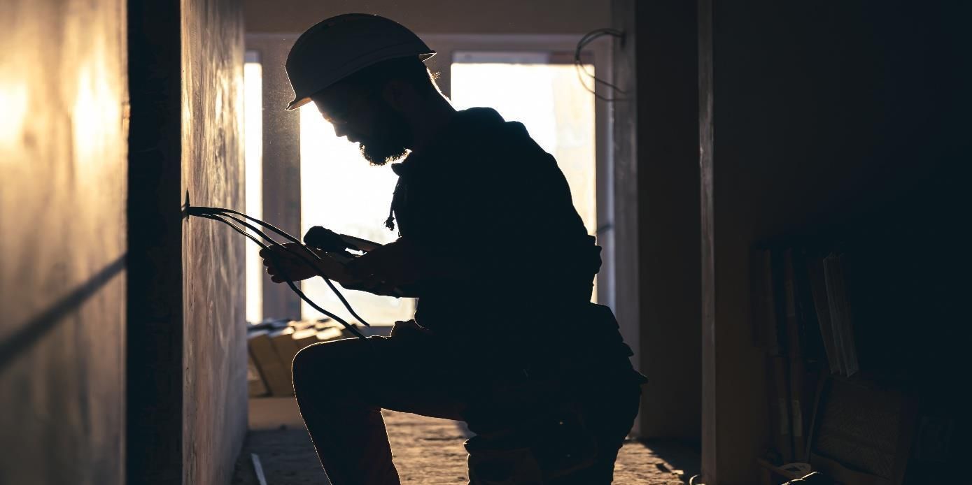 Silhouette of a worker in a hard hat using tools in a dim hallway lit from behind