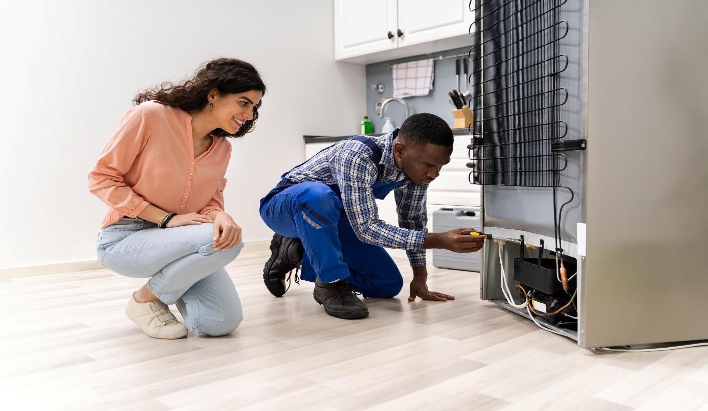 Two people crouch beside an open appliance in a bright kitchen, inspecting its exposed parts.