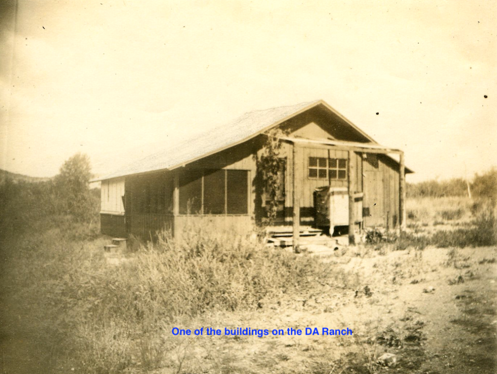 A weathered wooden building on a ranch with overgrown weeds.  The building has a screened-in porch.
