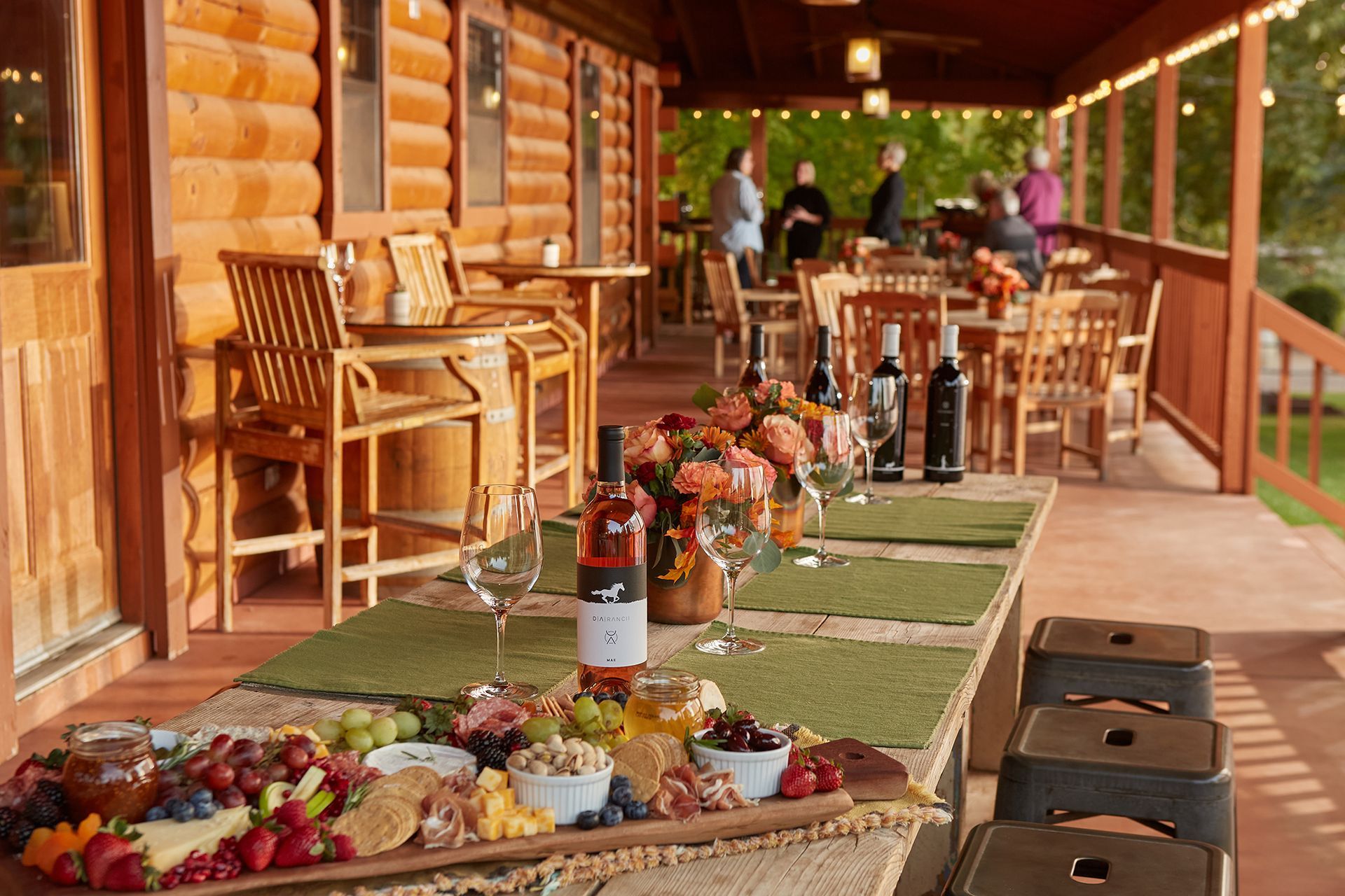 Outdoor patio with wine, charcuterie, and people near a log cabin.