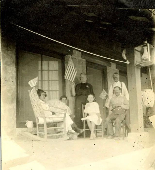Group of people on a porch, holding flags; identified: Mrs. Bates, Naquatsumi, Kent, Miss McClair, Eve, J.P. Girdner, William.