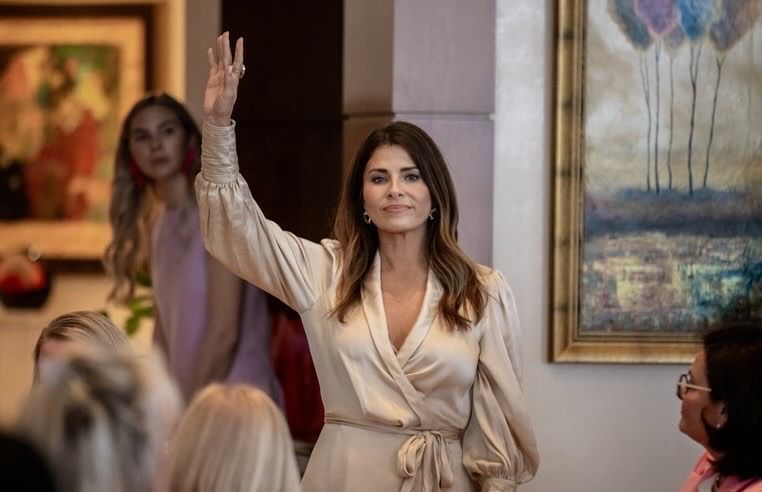 Woman in beige dress raises her hand at a formal indoor event, with attendees seated nearby.