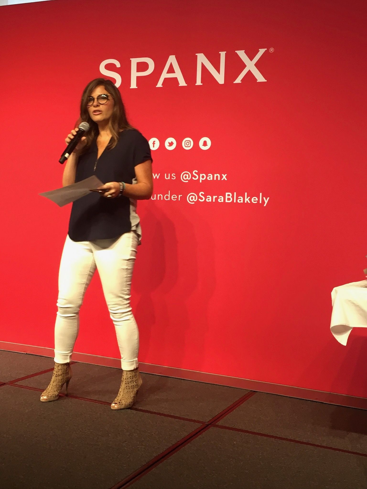 Woman speaking onstage at a SPANX event against a red backdrop, holding notes and a microphone.