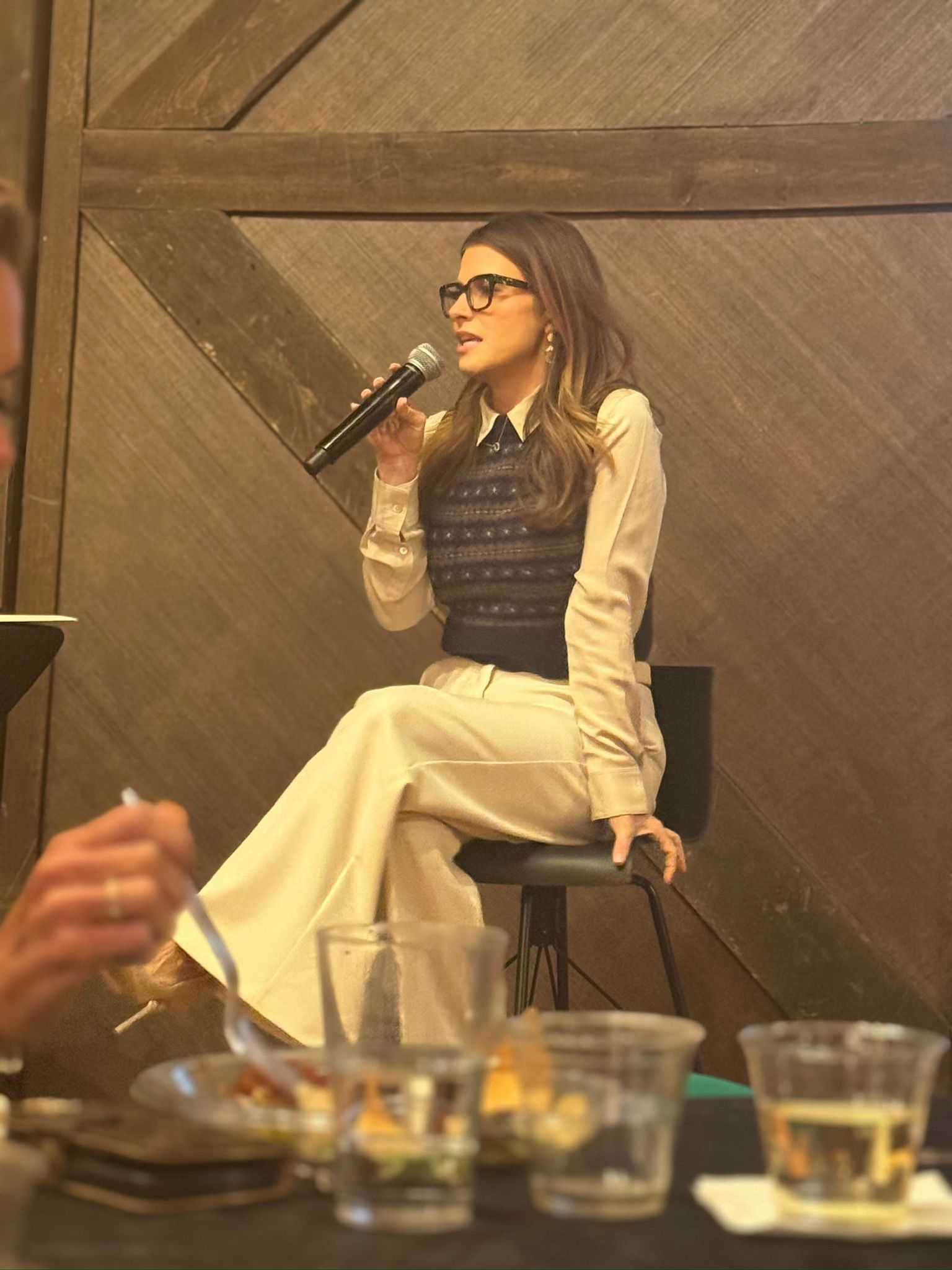 Woman singing into a microphone while seated on a stool in a warm-lit indoor venue.