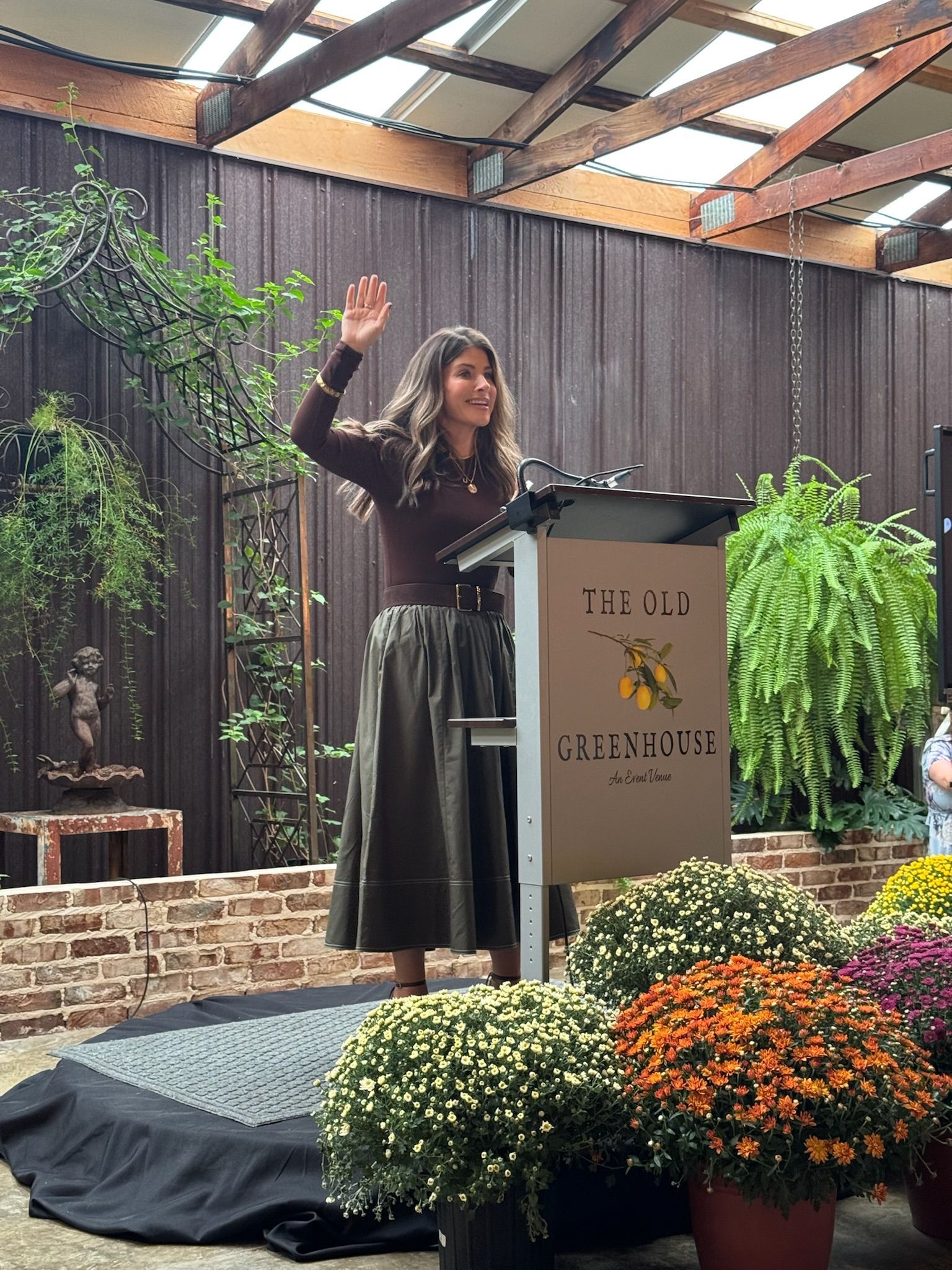 Speaker at podium on outdoor stage, raising hand amid flowers and greenery