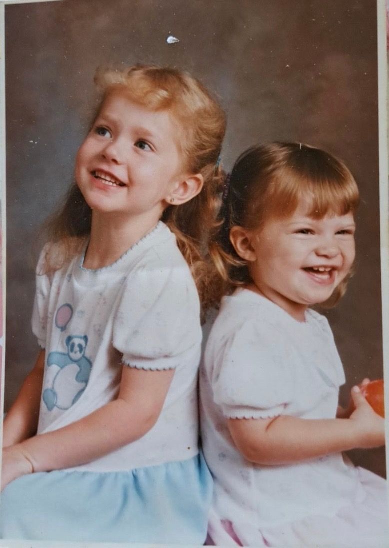 Two smiling children in white dresses sitting back to back against a studio backdrop