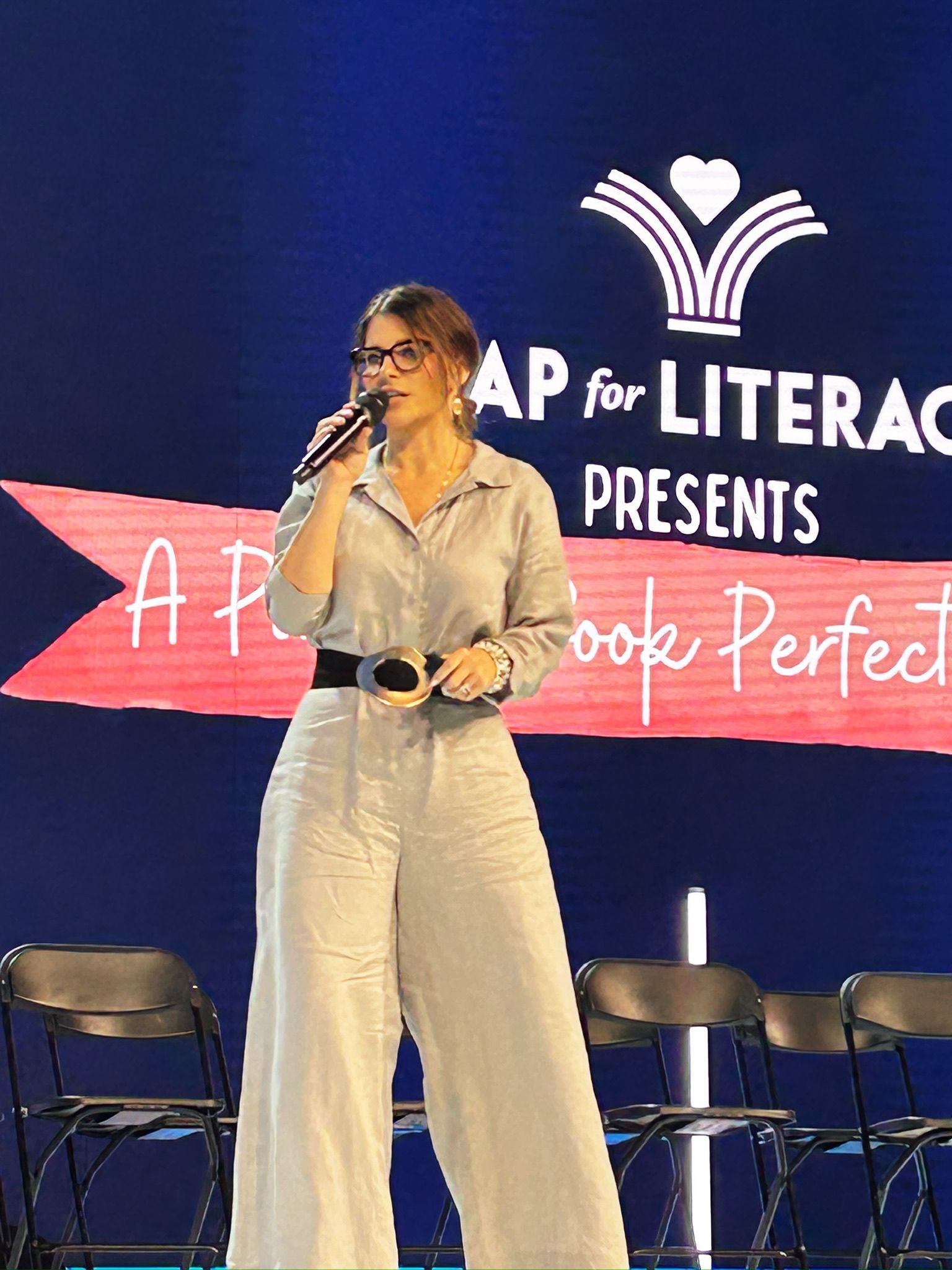 Woman singing on stage at AP for Literacy, wearing a light jumpsuit and sunglasses, with chairs behind her.
