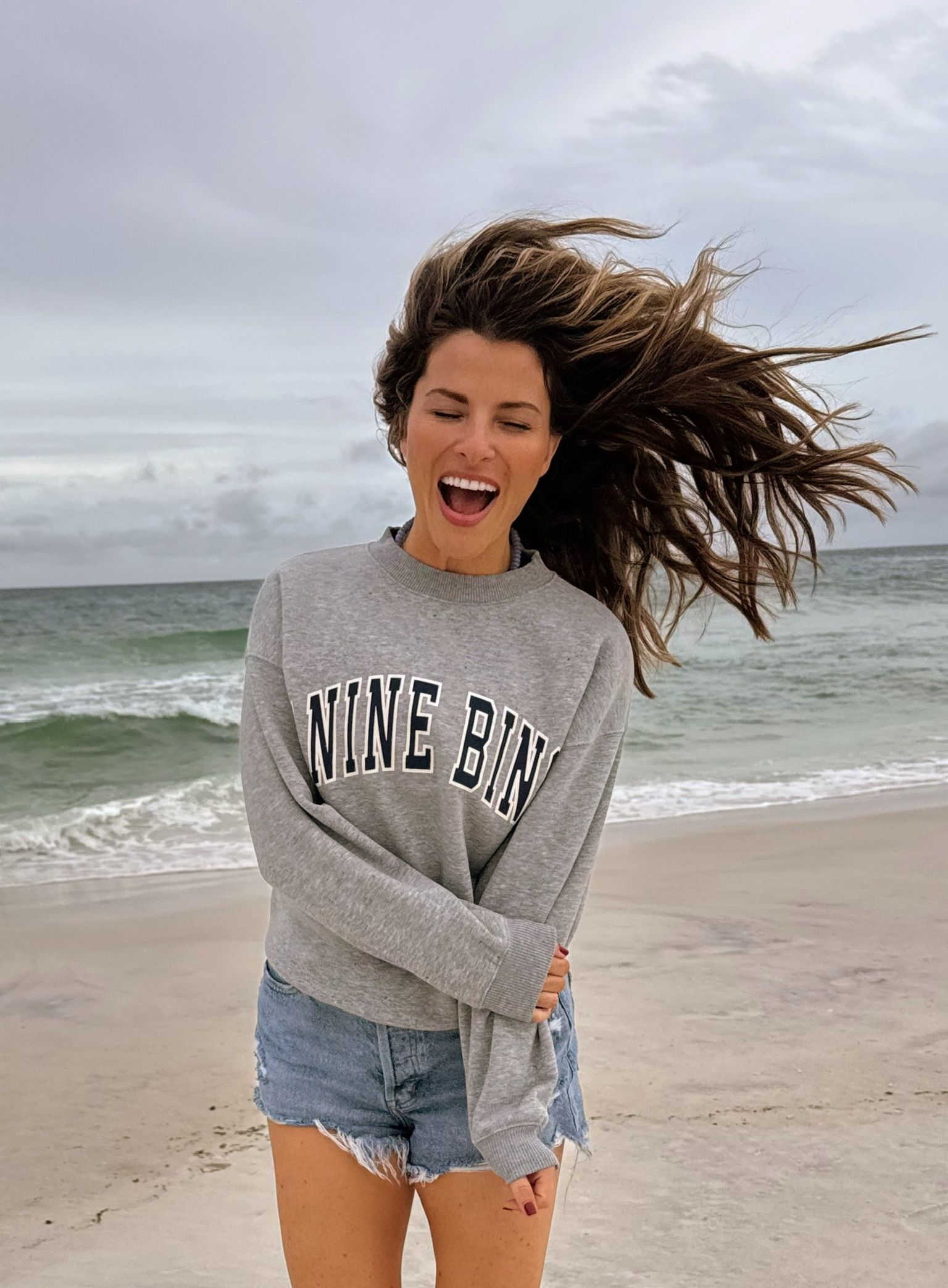 Woman on a windy beach wearing a gray sweatshirt and denim shorts, hair blowing across her face.