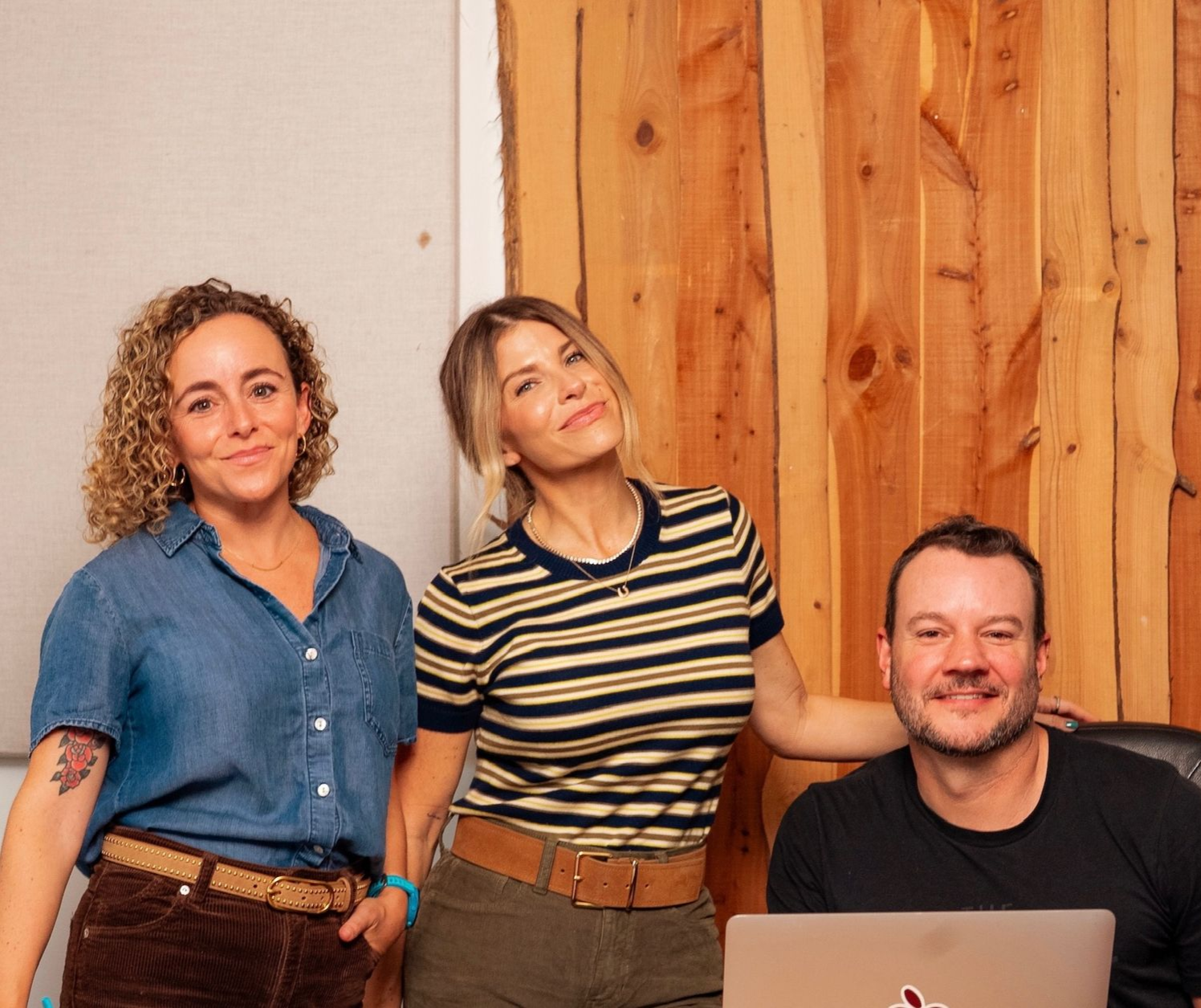 Three coworkers posing indoors beside a wooden wall, one seated with a laptop.