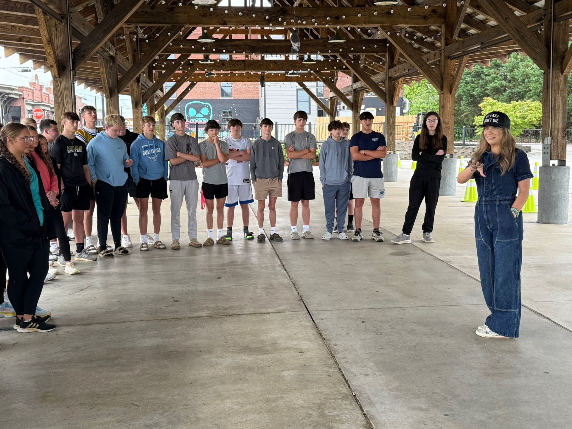 Group of people standing in a pavilion, listening to a woman speaking and gesturing during a gathering.