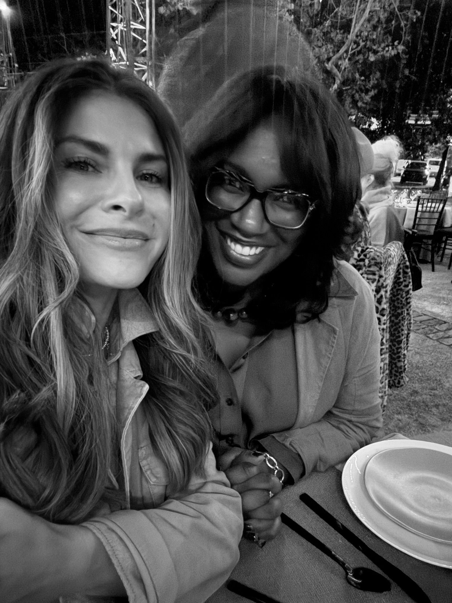 Two smiling friends at an outdoor café, posing closely beside a table with cups and utensils.