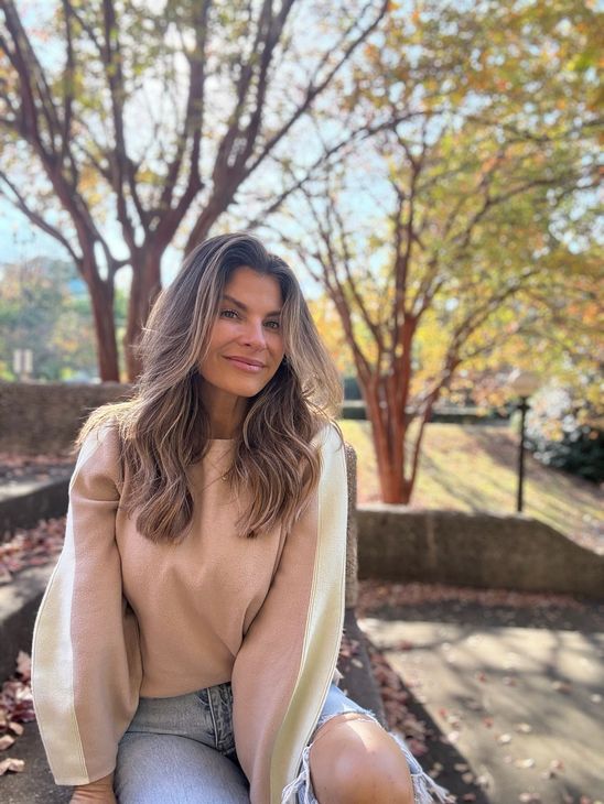Smiling person in a beige sweater and jeans sitting outdoors by trees in autumn light