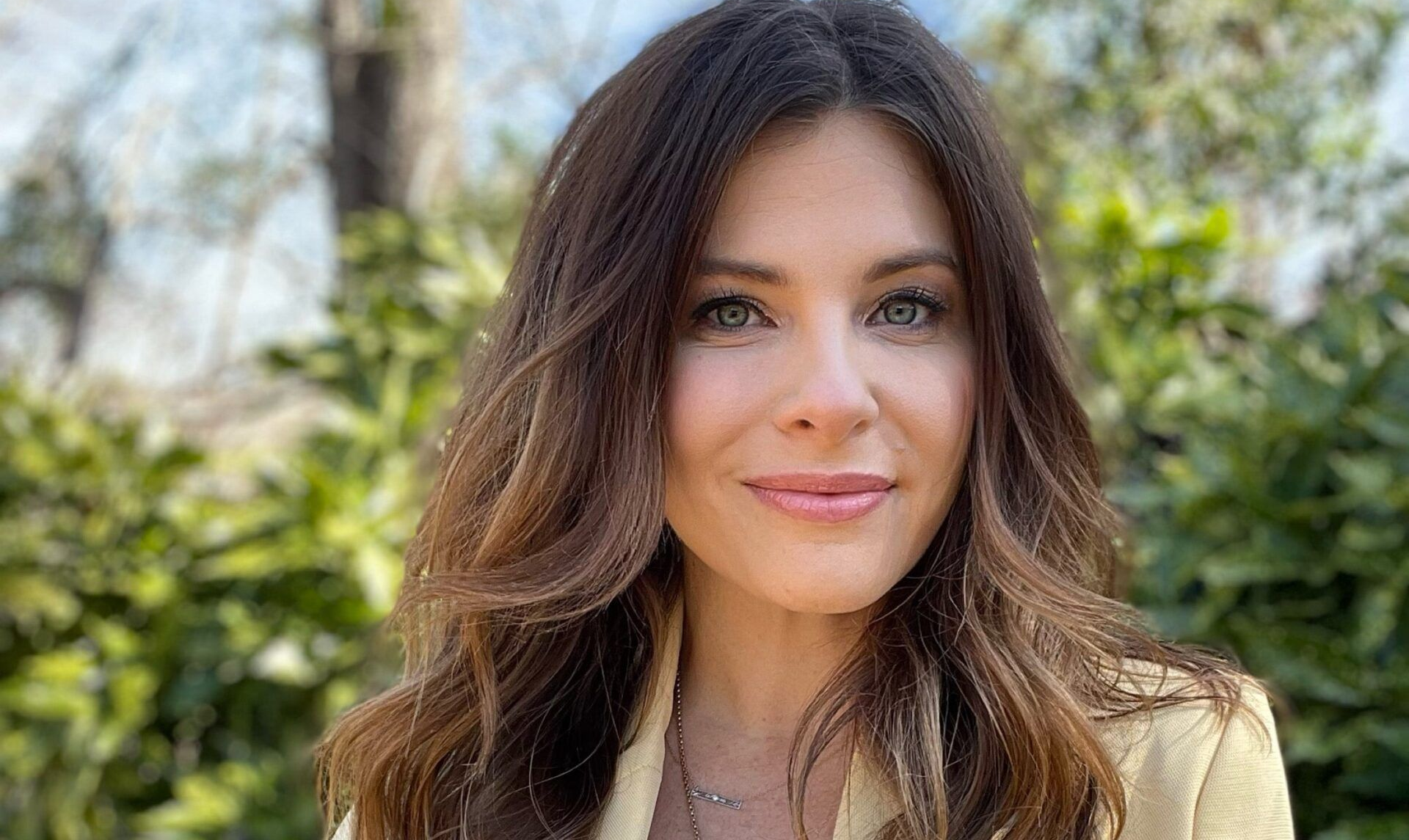 Smiling woman with long brown hair outdoors, with trees in the blurred background
