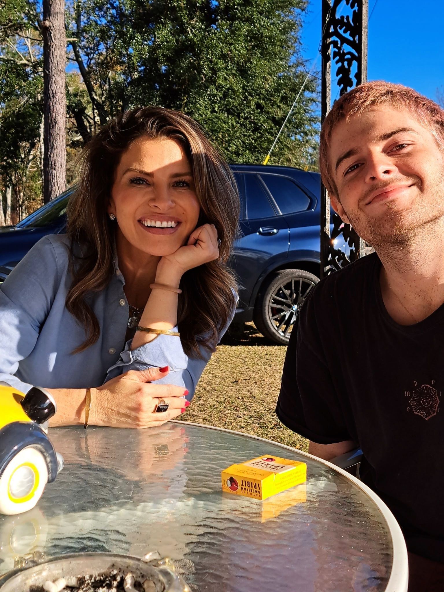 Two people smiling at an outdoor glass table in a sunny yard, with a toy truck and yellow box nearby.